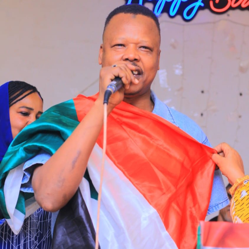 المطرب السوداني عامر عمر خلال إحيائه إحدى الحفلات بالقاهرة. (Photo: Private) رجل يحمل ميكروفونًا وجسمه مغطى بعلم ملون بالأبيض والأحمر والأخضر والأسود.