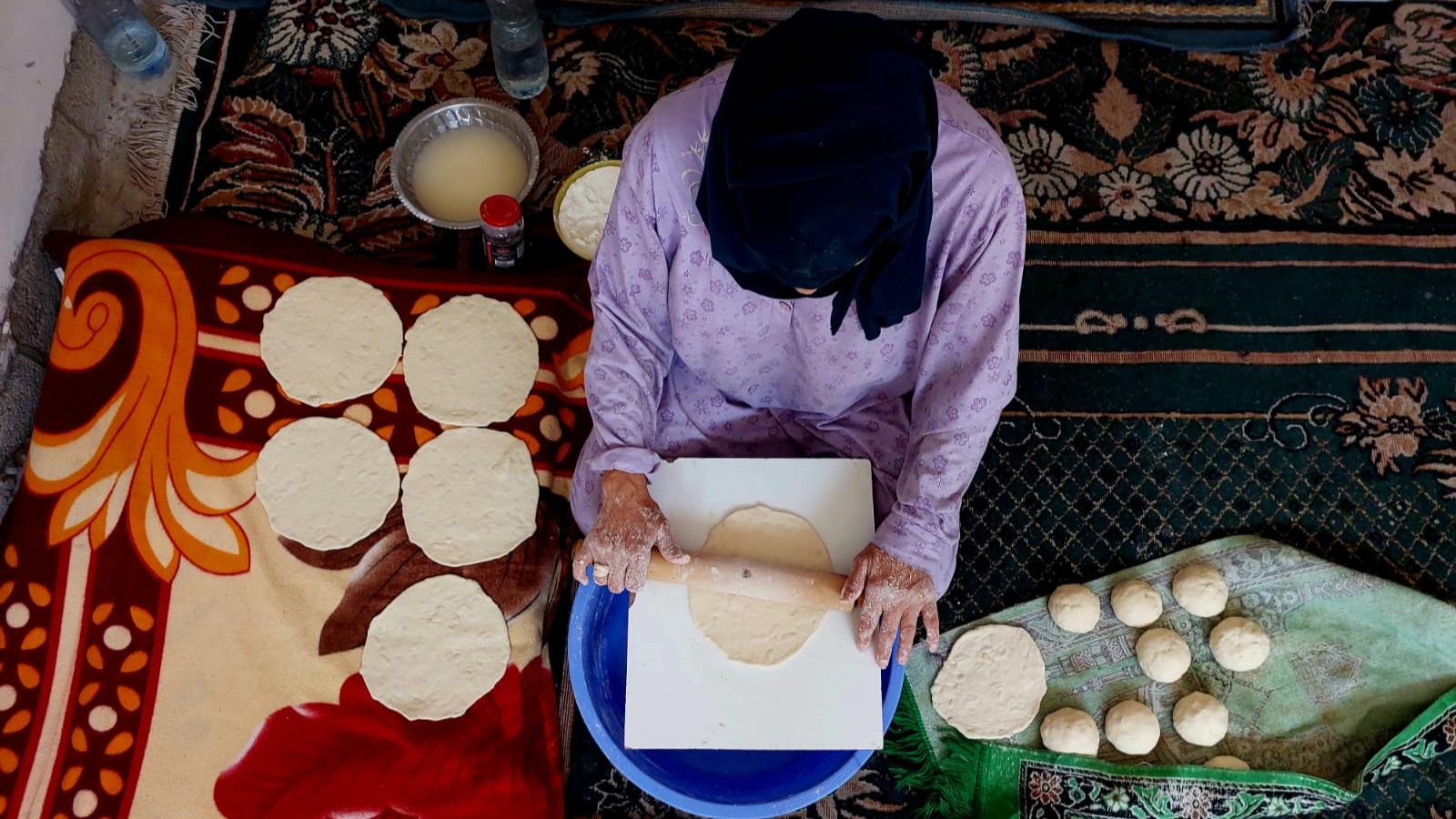 أم فلسطينية تخبز ما توفر من طحين داخل منزلها في مخيم النصيرات، 19 يونيو/حزيران 2025. (Photo: Copyrights for Qantara/Doaa Shahin) صورة من أعلى لإمراة تقوم بتحضير الخبز بمنزلها.
