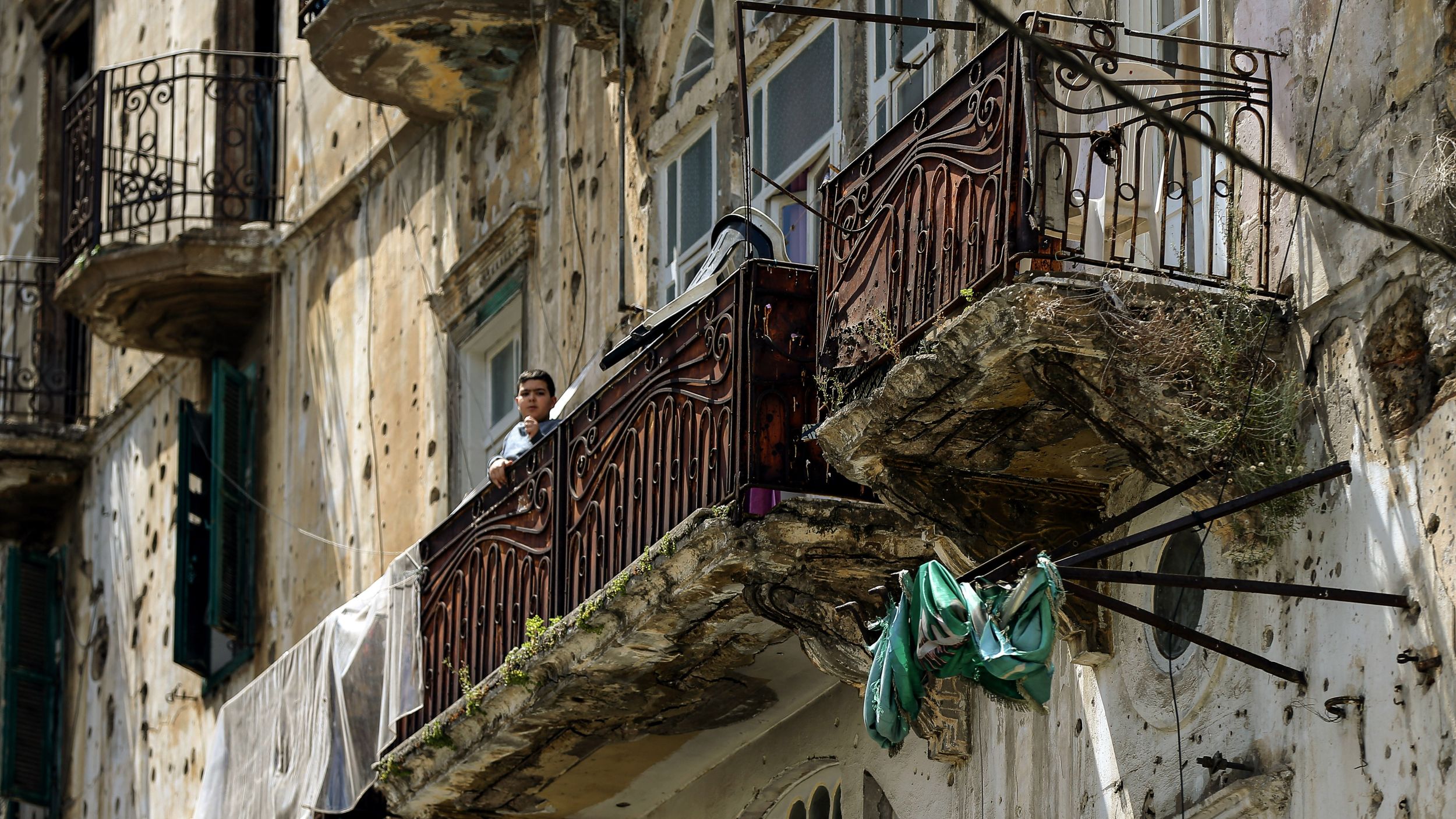 A boy looks out from the balcony of an apartment building marked with many bulletholes.