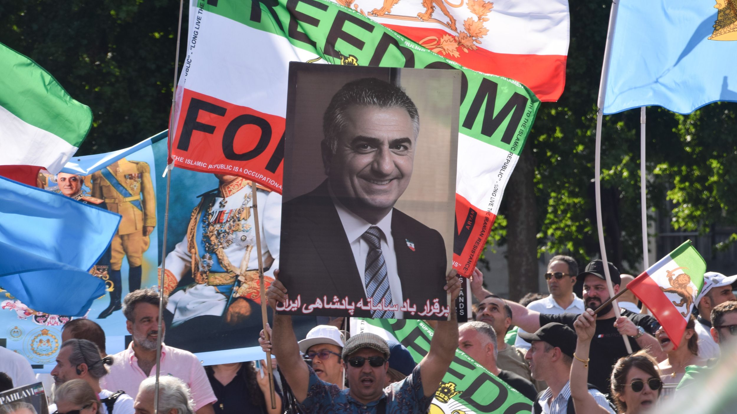 A man in a crowd holds up an image of Crown Prince Reza Pahlavi. There are many flags in the background, including the flag of Iran.