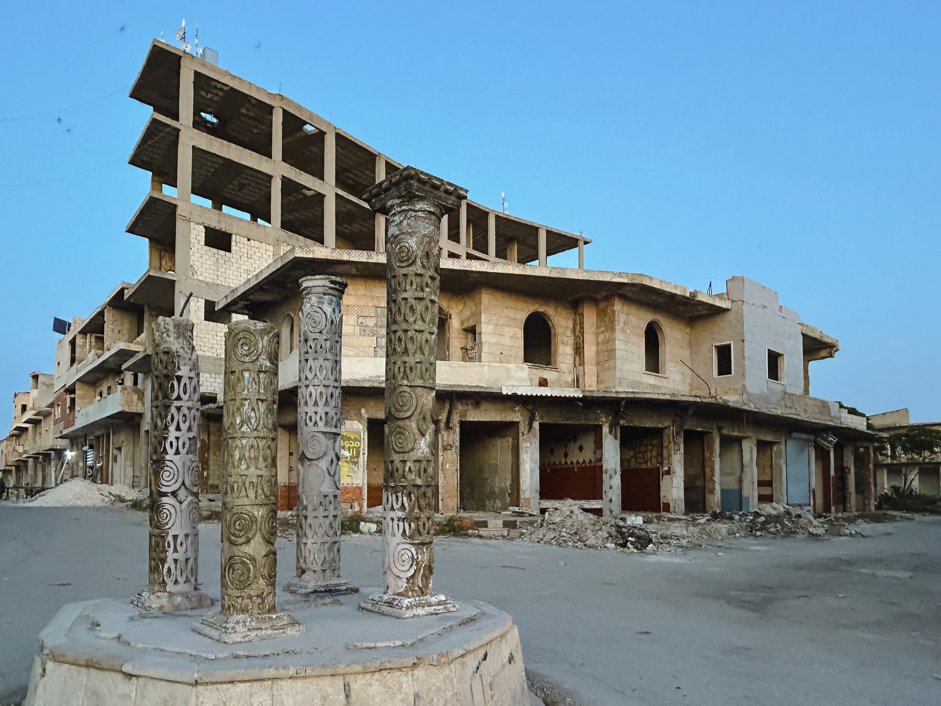 كفرنبل في أبريل/نيسان 2025. (Photo: Qantara/Kristin Helberg) مبنى متهدم، أمامه ثلاثة أعمدة تاريخية مزخرفة.