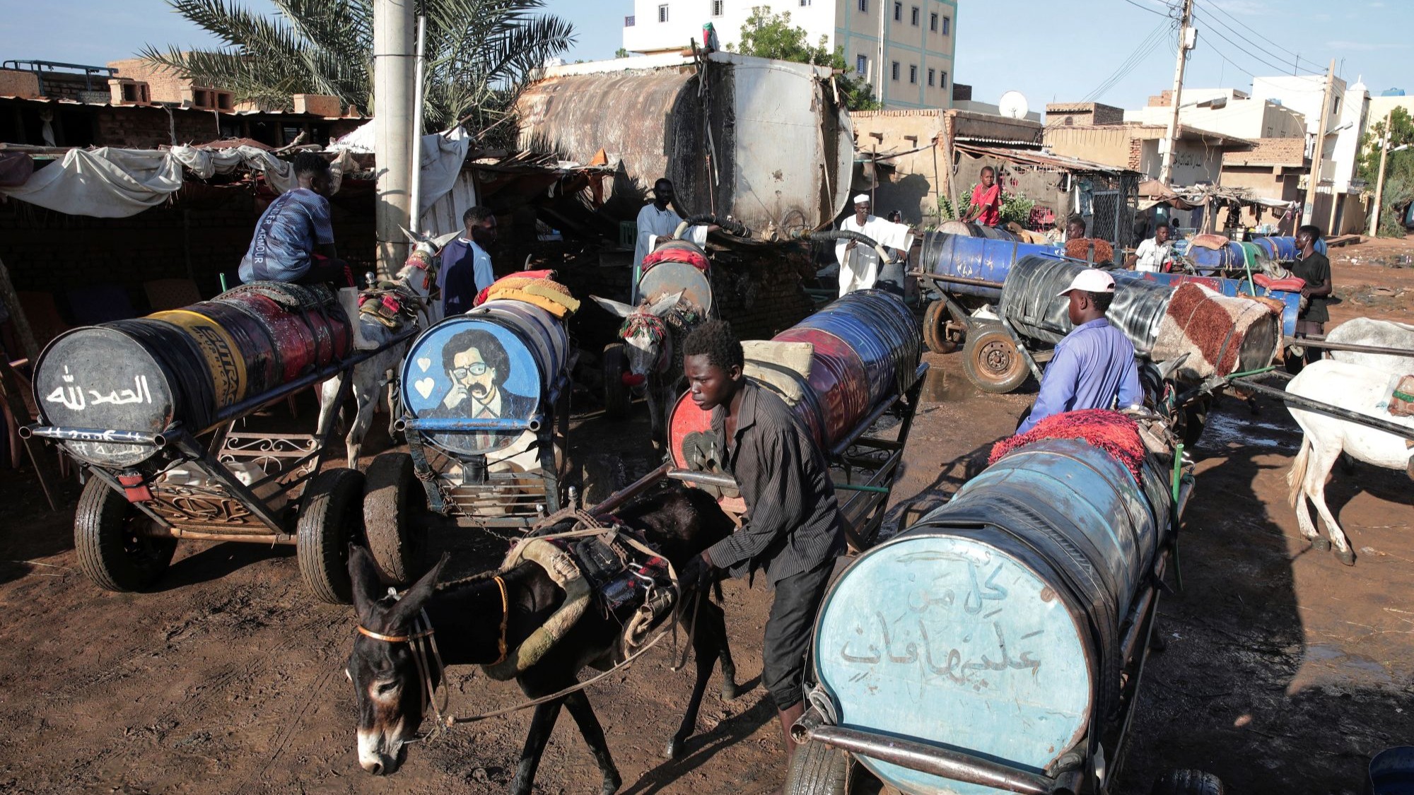 Donkeys pull carts with painted water barrels, some men stand by.