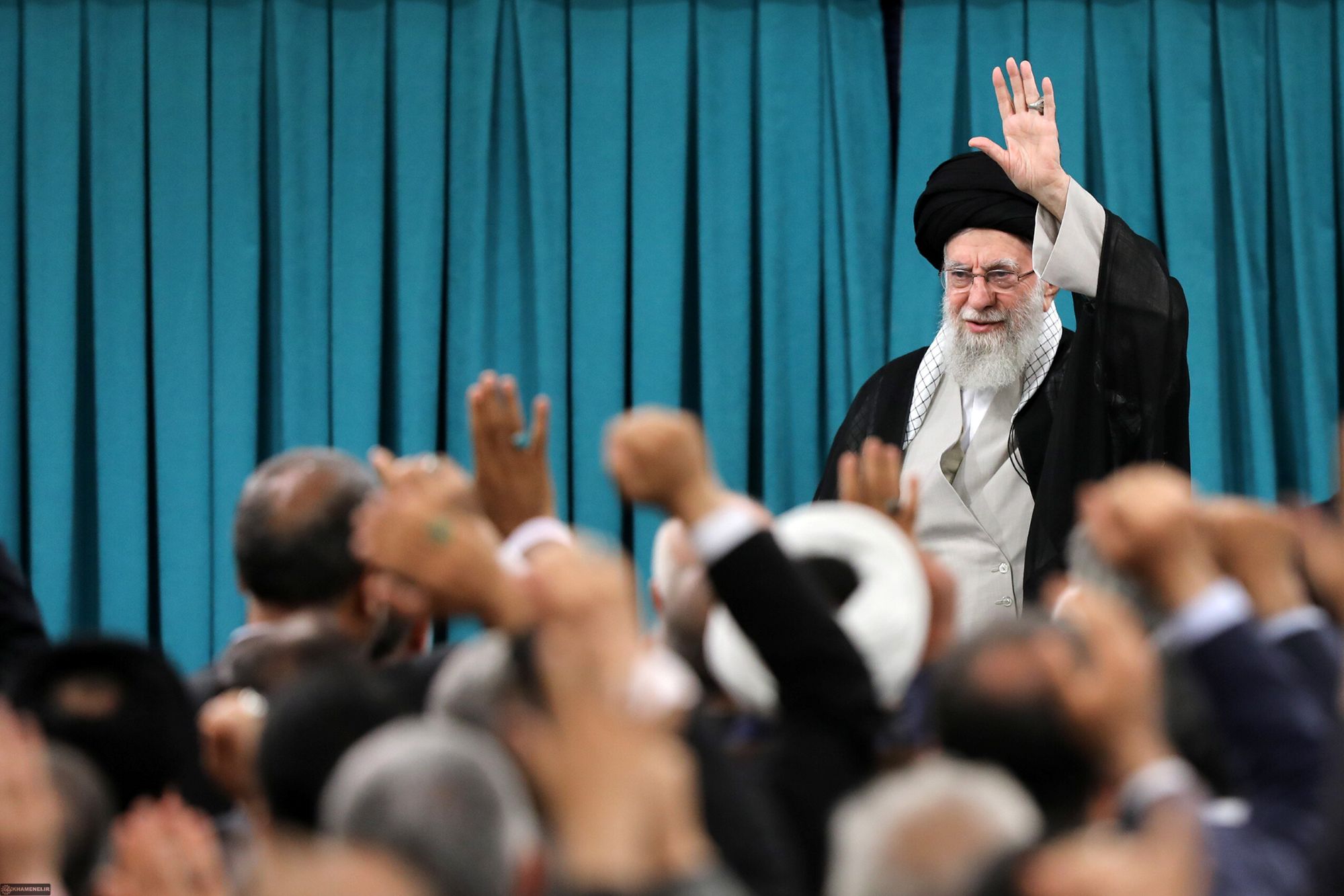 Ali Khamenei stands in front of a crowd of listeners and raises his left hand.