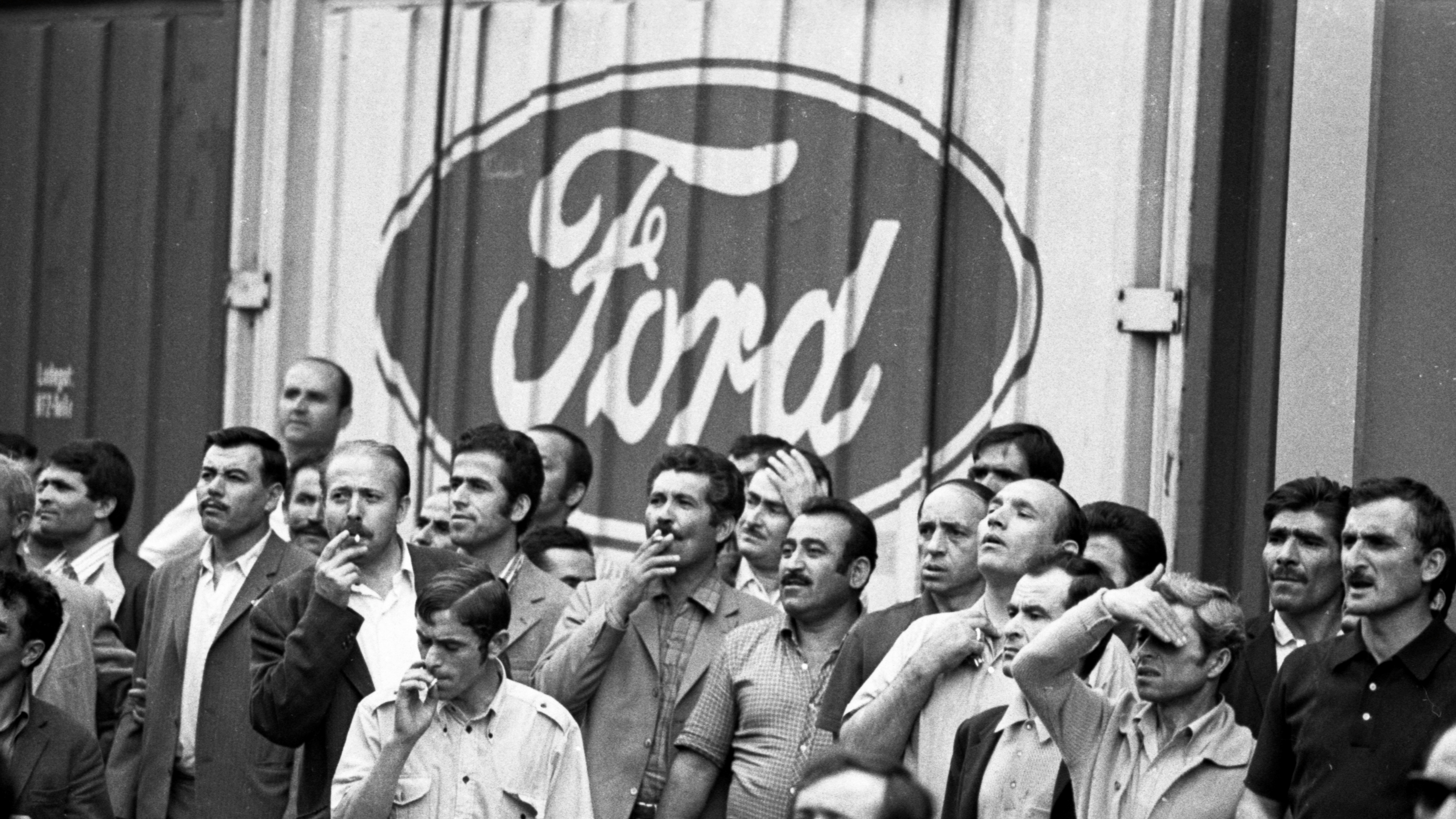 Schwarz-weiß Bild von Männern, die vor einer Wand stehen. Auf der Wand ist das Firmenlogo von Ford zu sehen.