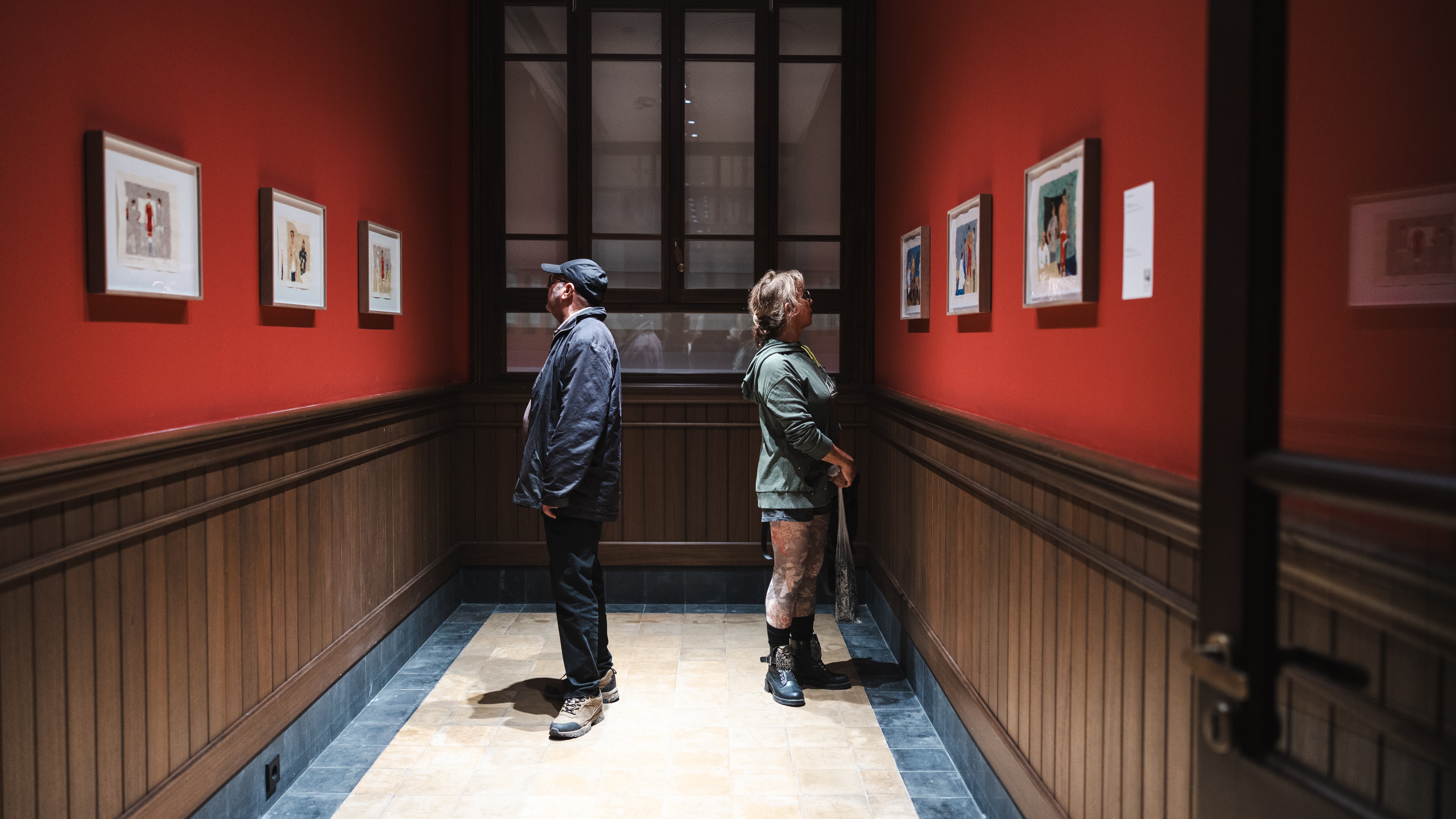 18th Istanbul Biennial (Photo: IKSV | Mete Kaan Özdilek) Two people in a corridor, facing away from eachother and looking at paintings hanging on the walls.