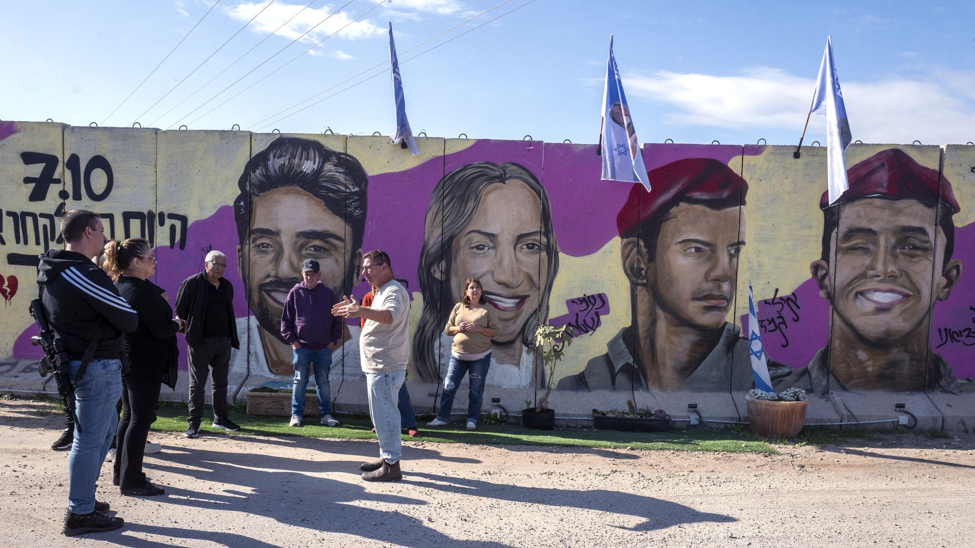 Eine Gruppe steht vor einer Betonmauer. Auf der Mauer sind farbige Portraits von israelischen Geiseln zu sehen.