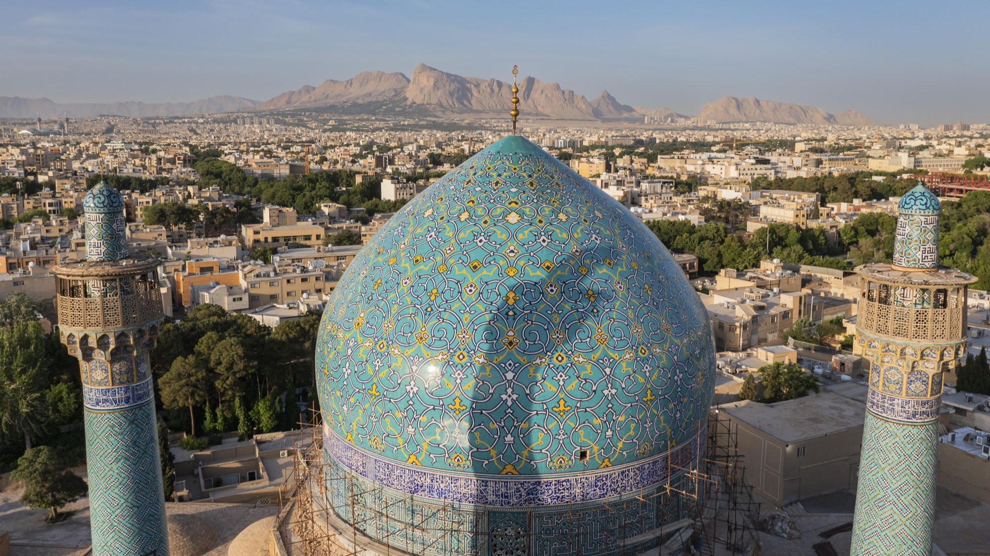 Sheikh Lotfollah Mosque in Isfahan, Iran. (Photo: Picture Alliance / Amazing Aerial Agency | Airpano) A mosque dome with green mosaic tiles against the backdrop of a city, with mountains in the background.