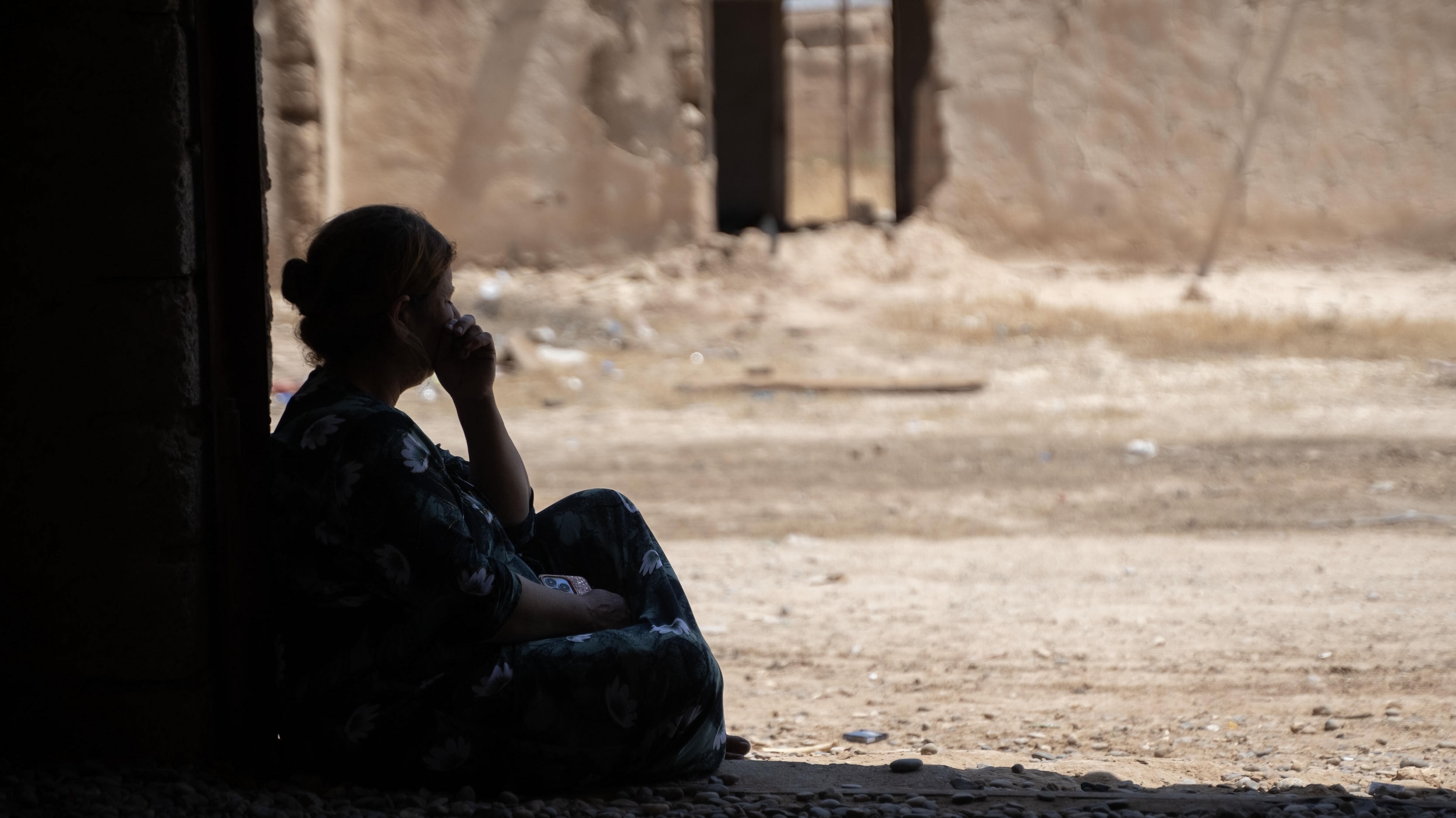 A silhouetted woman sits in the doorway of a ruined building looking out onto the street.