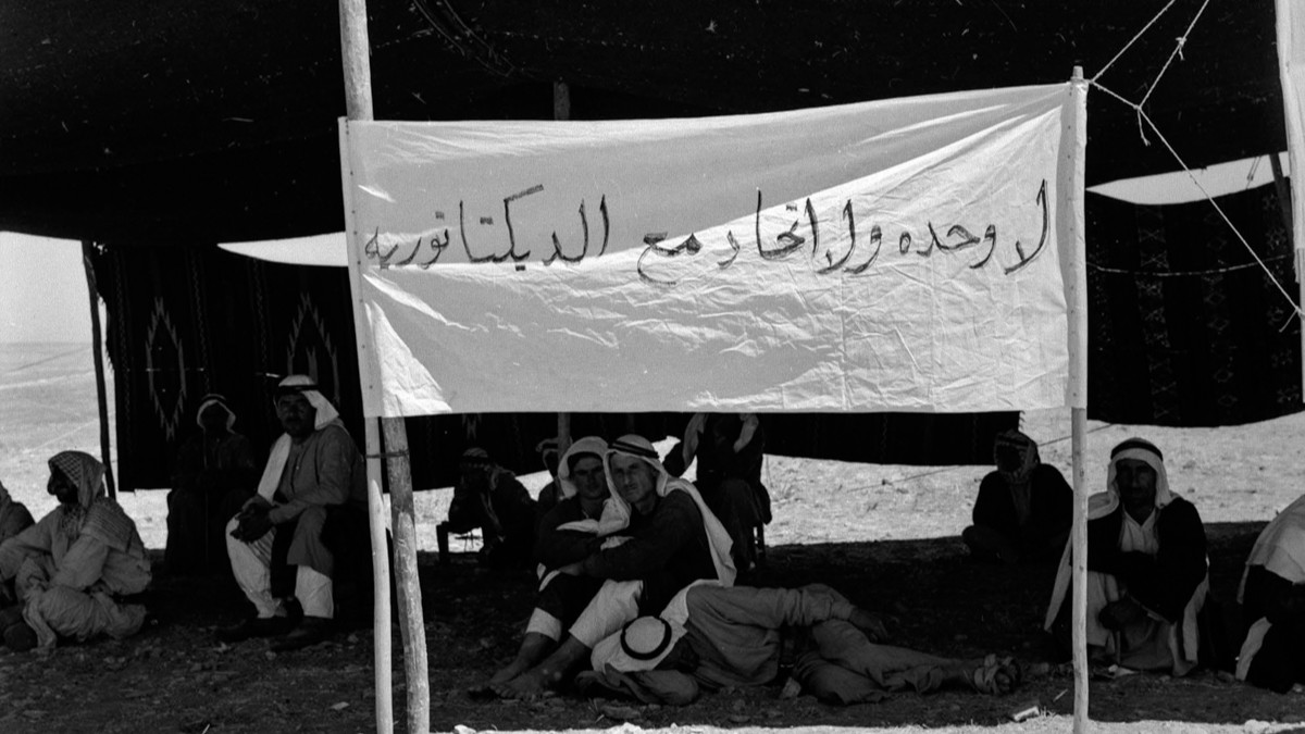 Schwarz-weiß Bild: Männer ruhen im Schatten, vor ihnen ist ein Transparent aufgespannt.