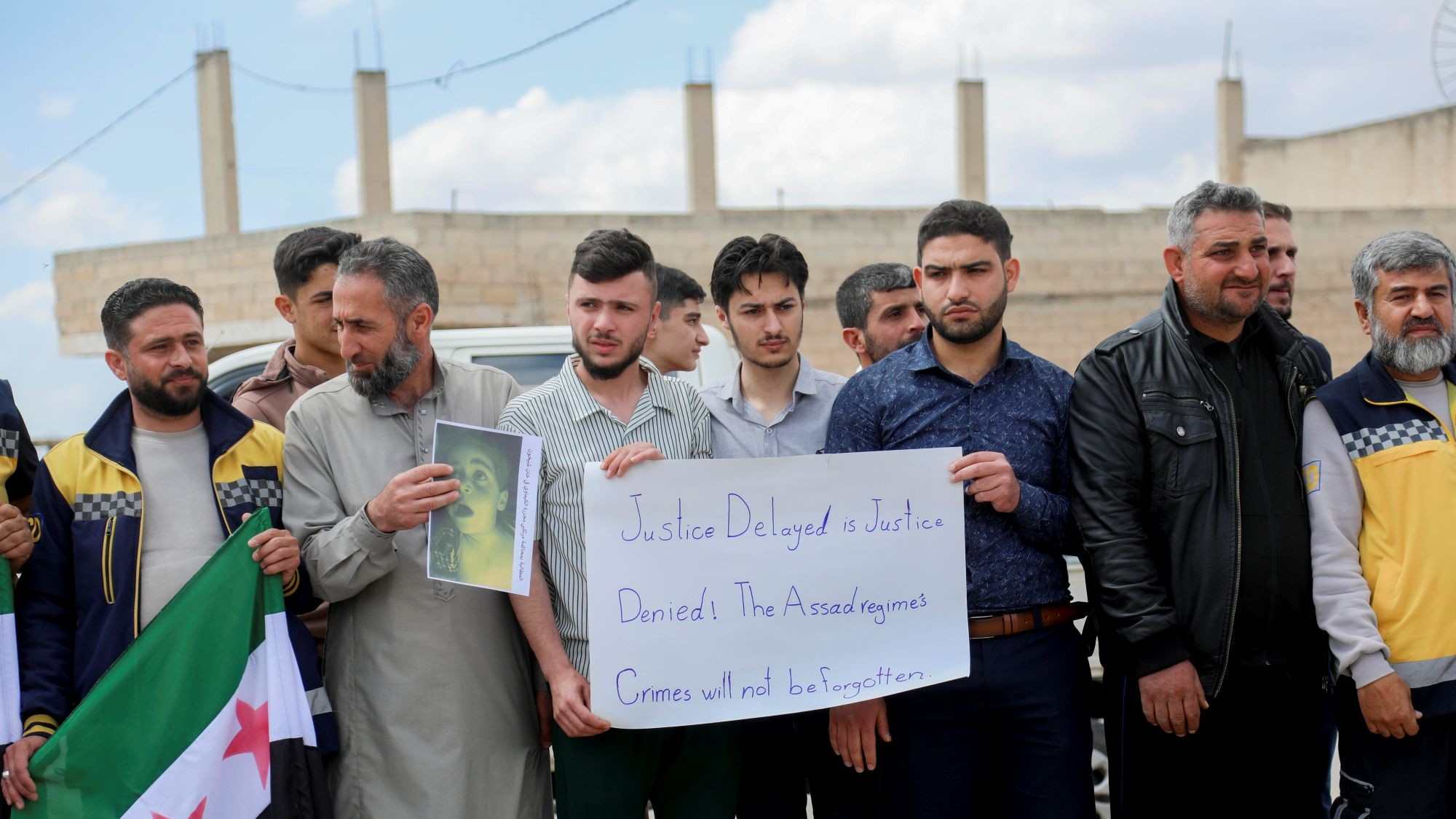 Eine Gruppe Männer hält Plakate und die neue syrische Flagge. Plakat: "Justice Delayed is Justice Denied! The Assad regime's crimes will not be forgotten."