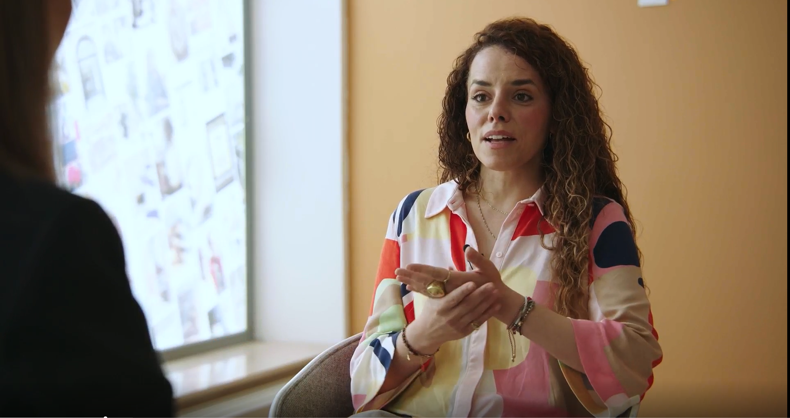 Eine Frau in bunter Bluse sitzt vor einem Fenster und spricht mit ihrem Gegenüber.