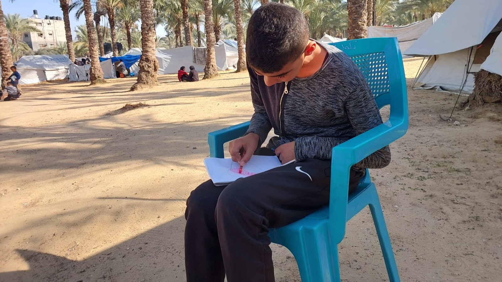 فتى يجلس على كرسي وبيده قلم وورقة، وفي الخلفية تظهر خيام وأشجار النخيل. (Photo: Qantara/Doaa Shaheen)