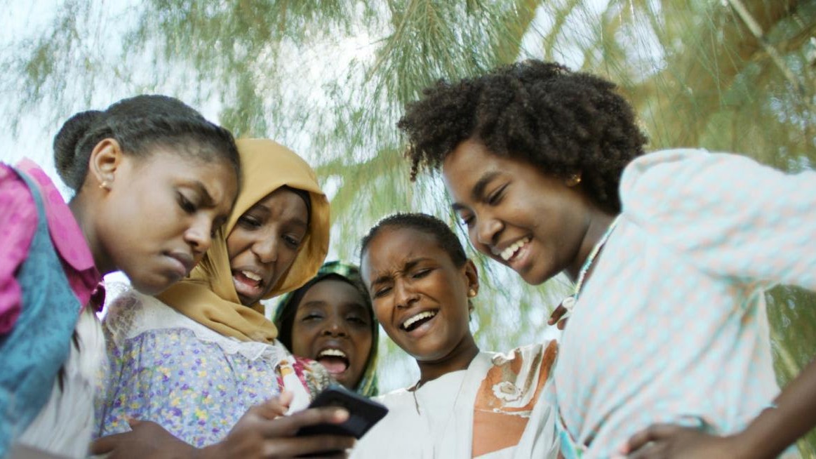 Five girls look at a mobile phone together and react with different emotions.