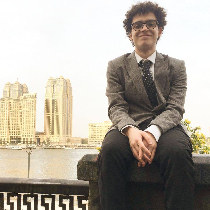 شاب يرتدي نظارة ويجلس على حافة كوبري وخلفه مجرى مياه، ومبان شاهقة