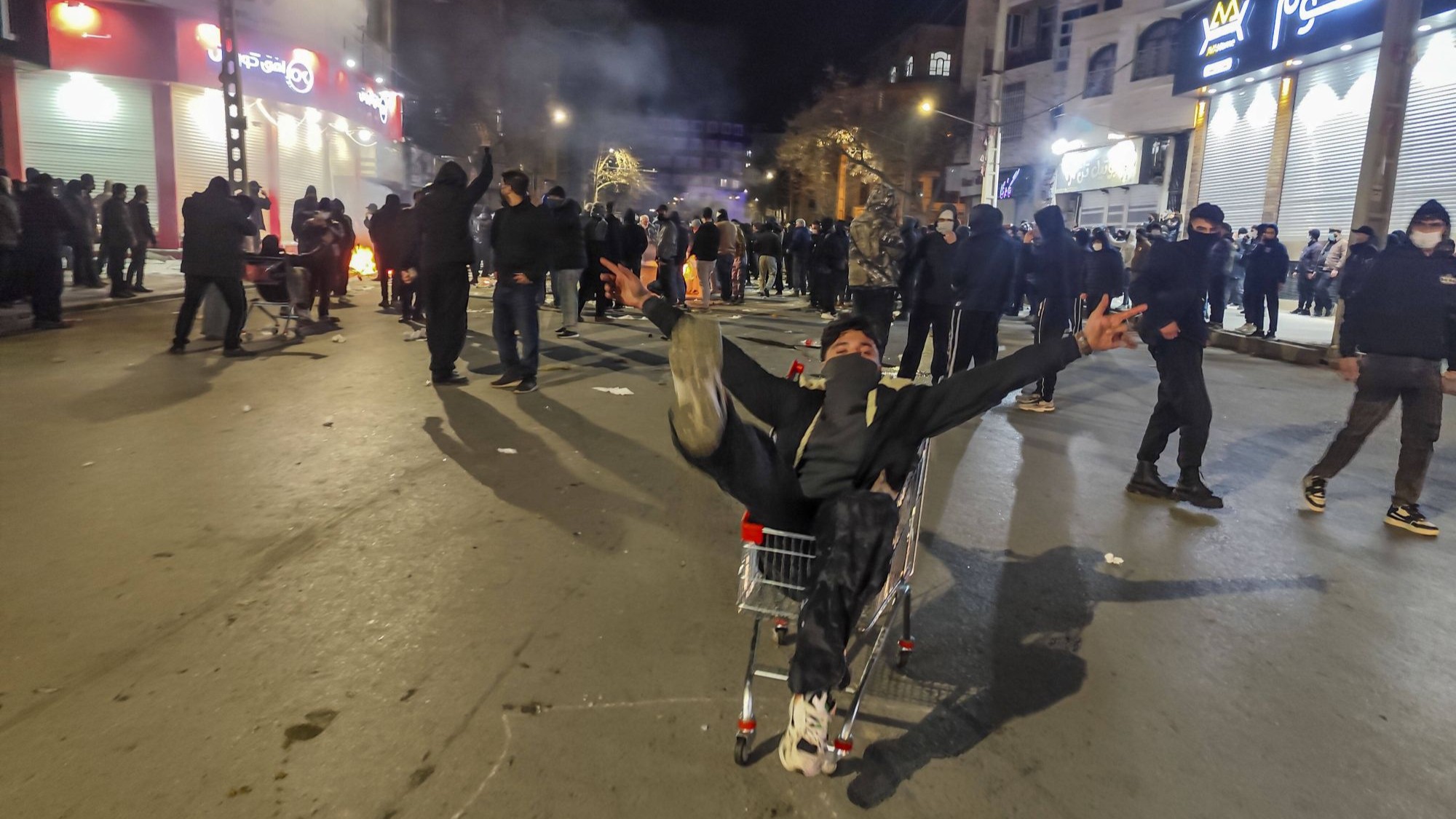 A man wearing a lower face covering sitting in a shopping trolley. Around him are other people protesting.