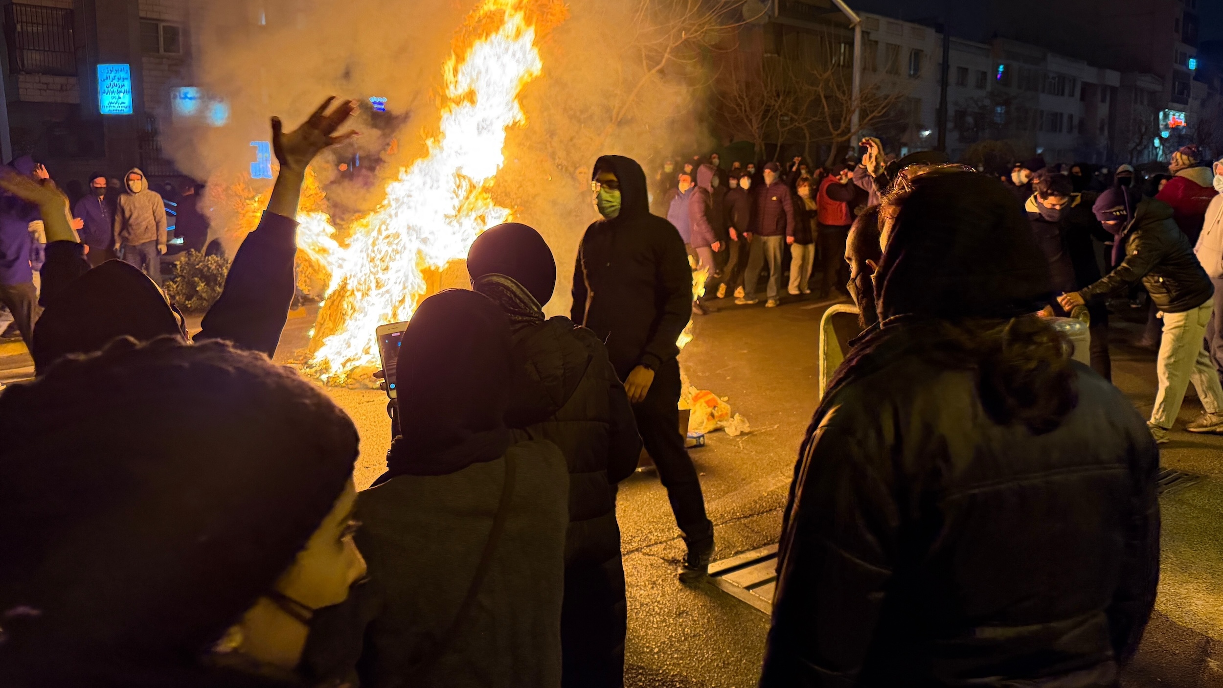 A crowd of people surround a fire in a street at night.