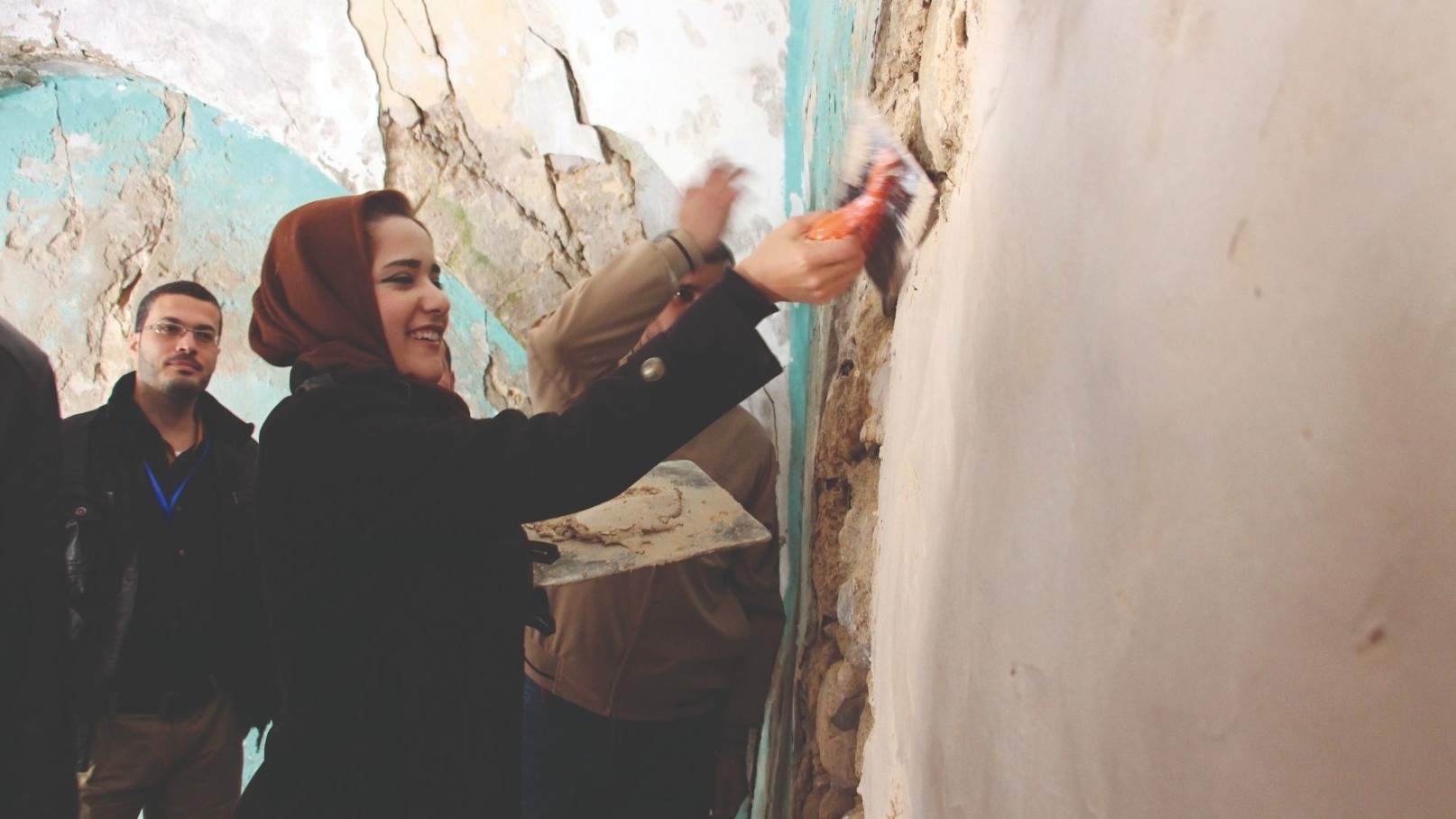 A young woman is holding a wall repair brush, and behind her are two young men.