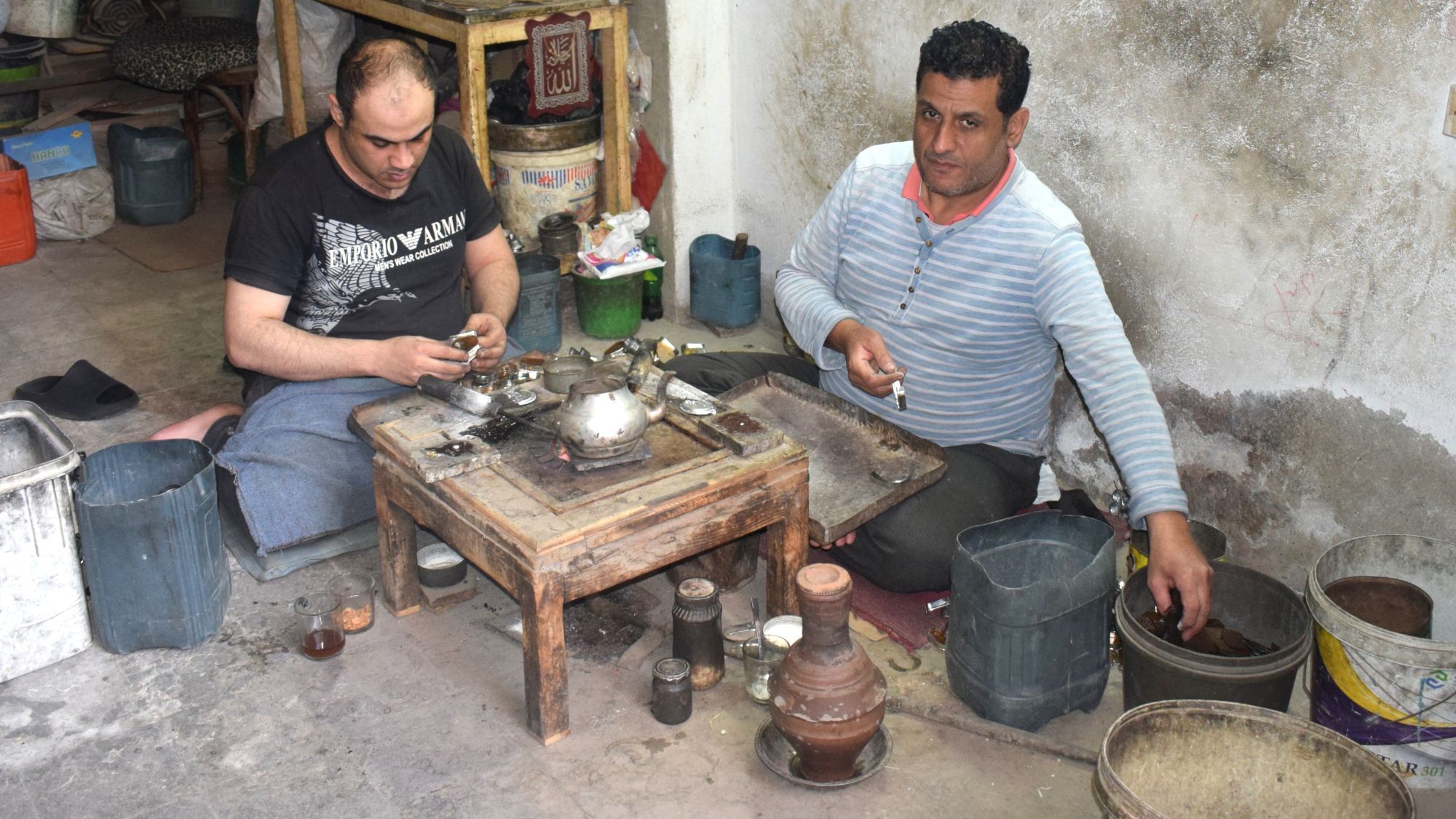 رجلان يجلسان أمام منضدة تبدو كورشة بدائية. 