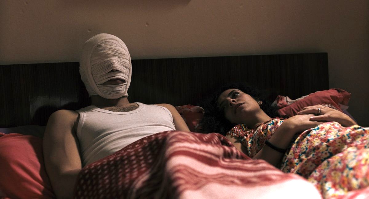 Ein Paar liegt im Bett, der Mann trägt eine Bandage um den Kopf und das Gesicht.