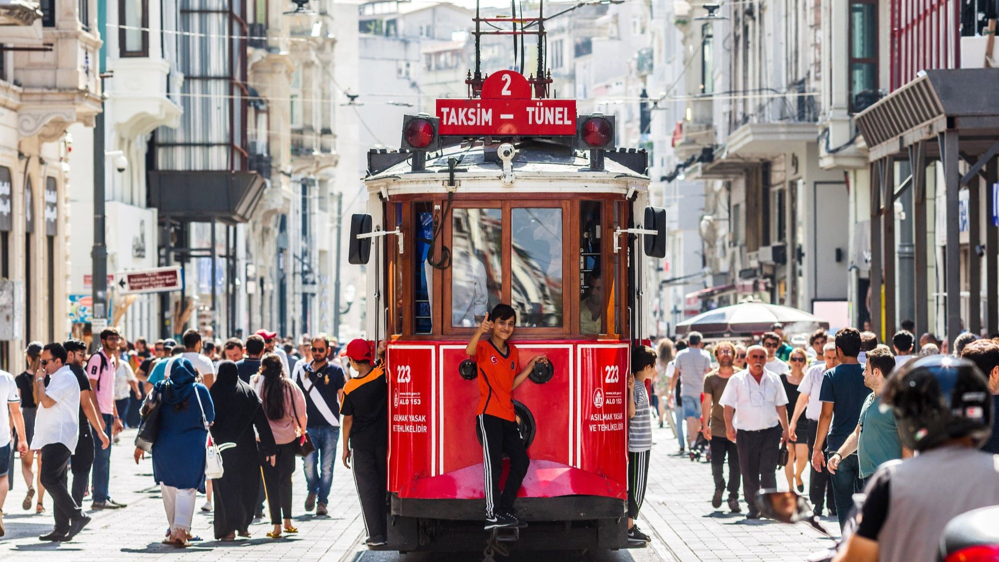 Ein kleiner Junge zeigt den Daumen nach oben, er fährt hinten auf einer roten Tram durch eine belebte Straße.