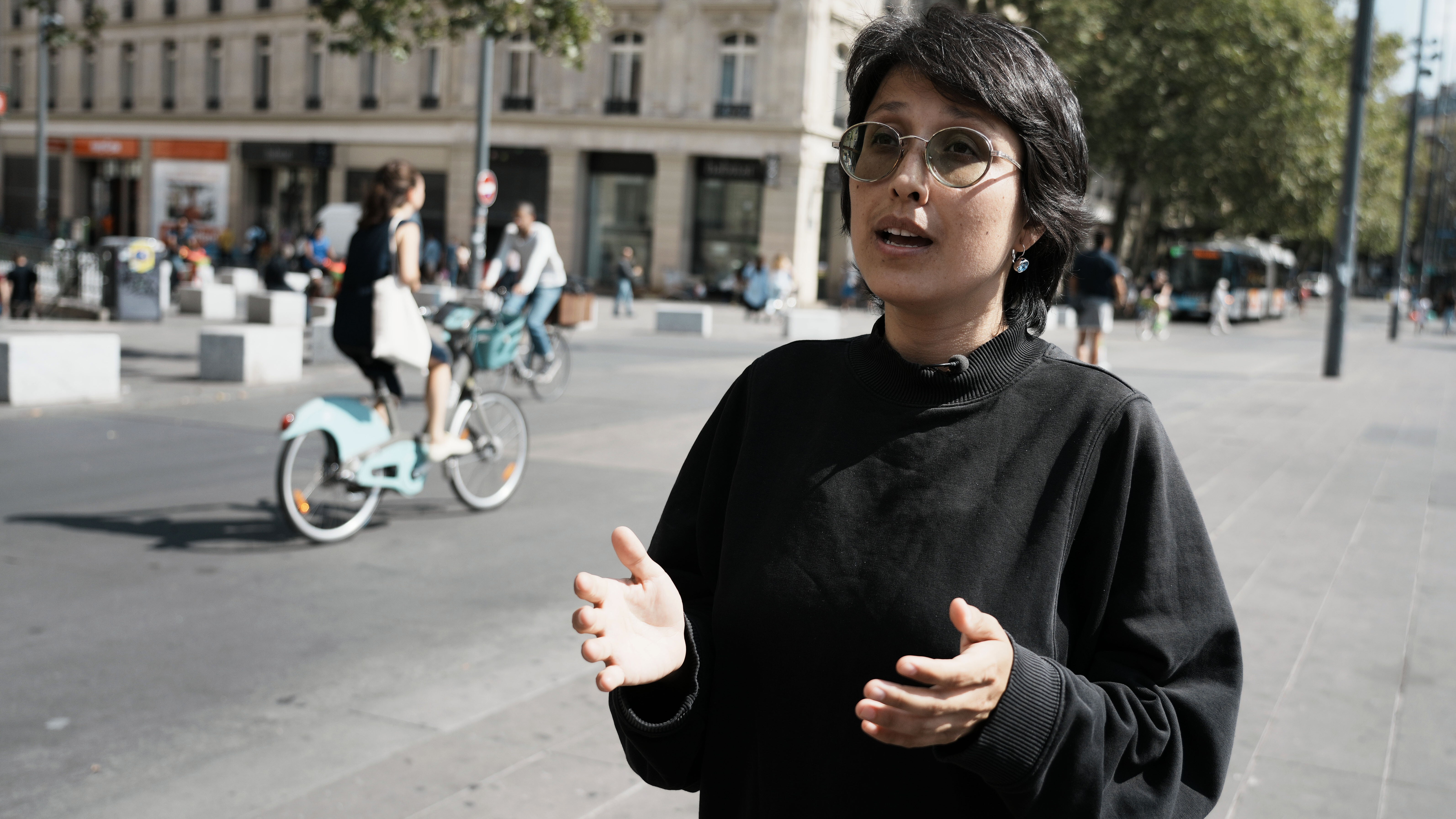 Shahrbanoo Sadat gestikuliert. Im Hintergrund eine Straßenszene, links im BIld eine Fahradfahrerin. 