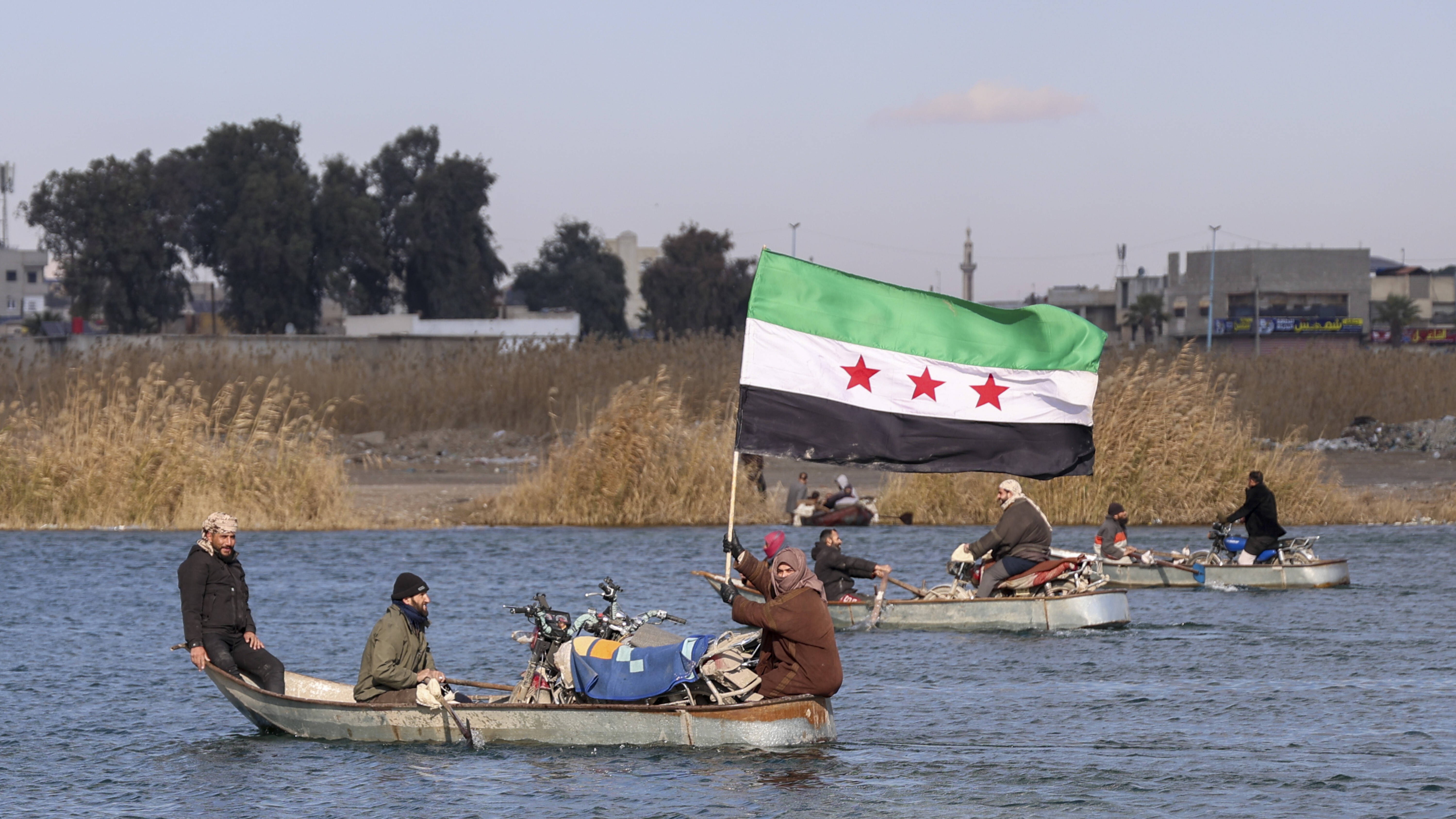 Ein Boot fährt über einen Fluss. Eine Passagierin hält die syrische Nationalflagge in den Händen. 