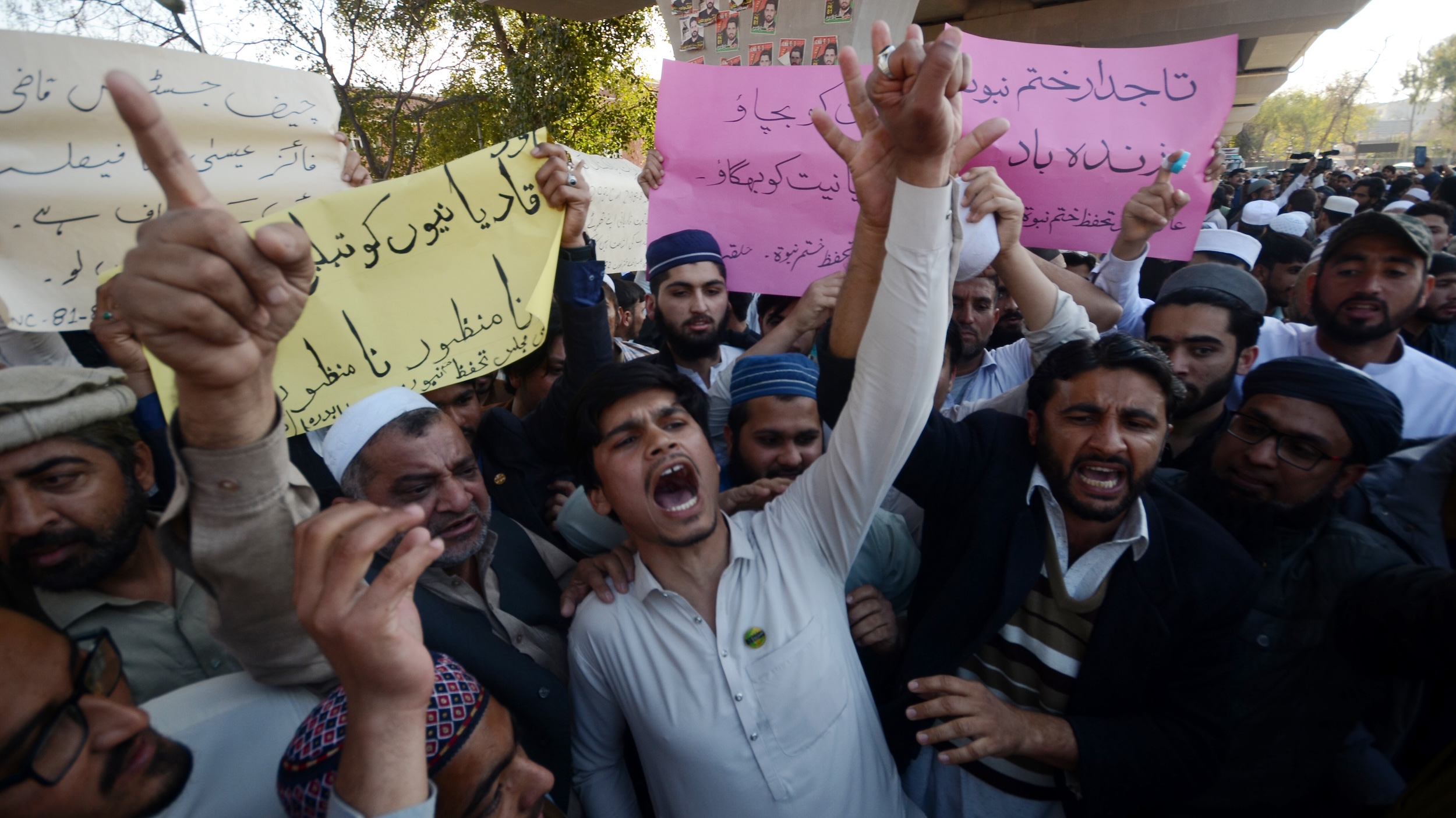 A group of men shouting with hands raised. Some hold signs with Urdu text.