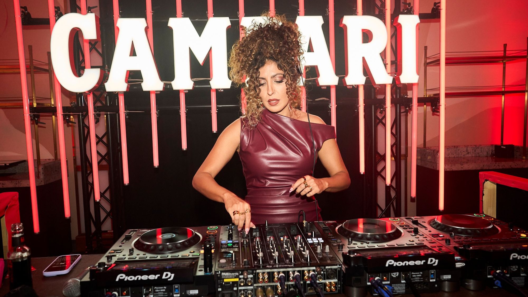 Eine Frau in dunkelrotem Kleid und hochgesteckten Haaren steht am DJ Pult. Hinter ihr Leuchtreklame.