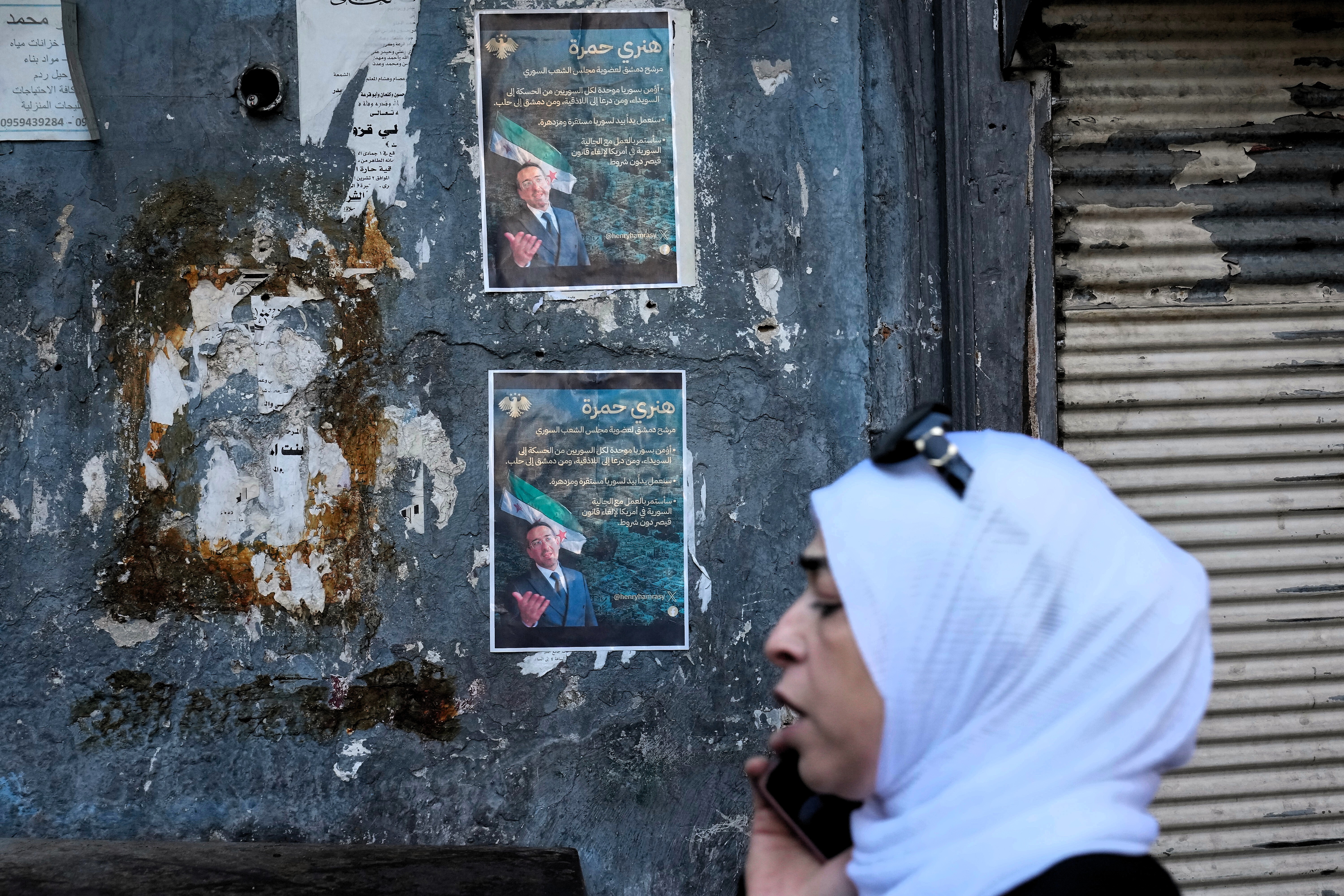 A Syrian woman passes in front of posters showing Syrian-American Jew Henry Hamra, a candidate for the Syrian Parliamentary elections, in the Jewish neighborhood of old Damascus, Syria Friday, Oct. 3, 2025.