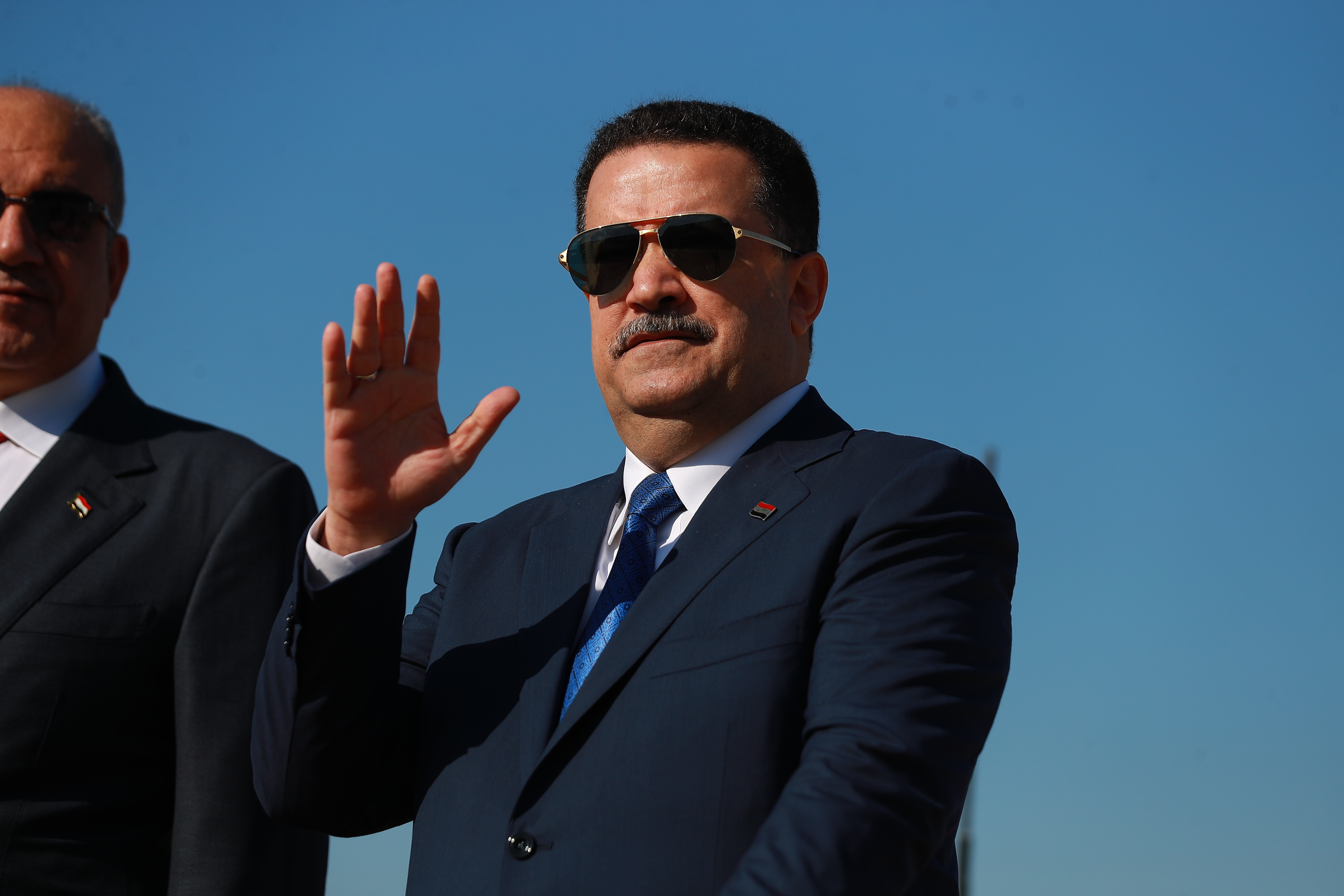 A man in a suit and sunglasses raises his hand to wave at people.