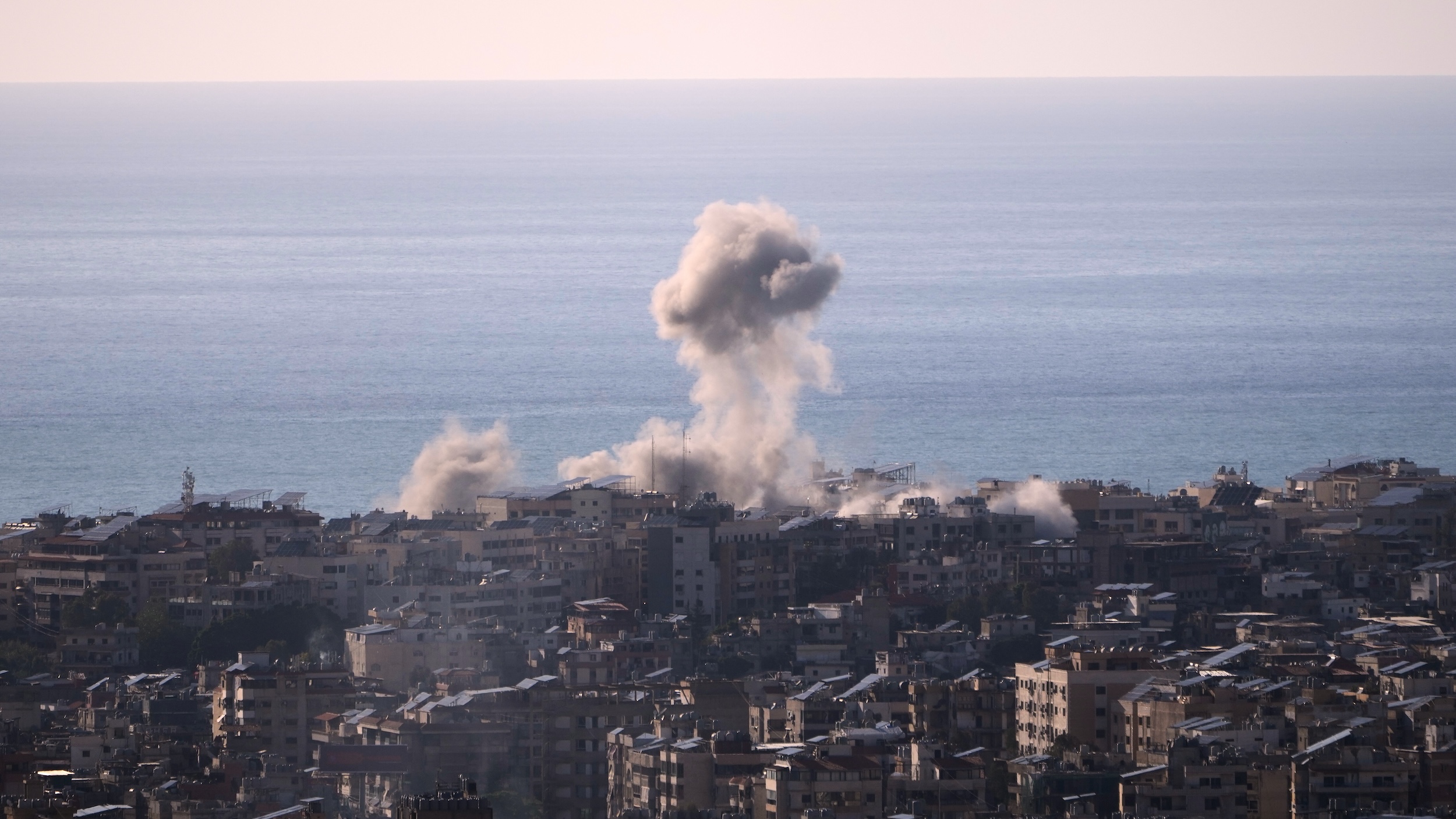 A plume of smoke rises from a building in a dense Beirut neighbourhood. The sea and the sky are in the background.