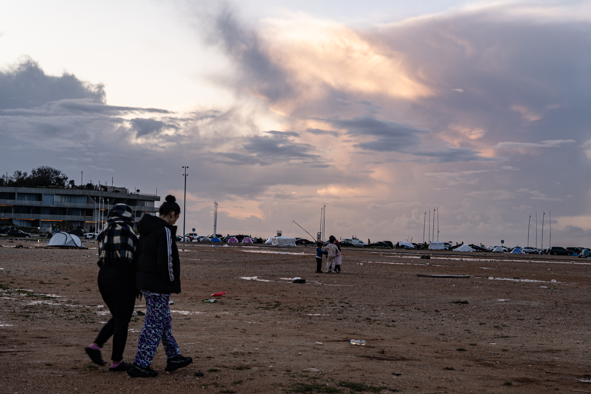 Two people walk across an empty lot, tents are pitched in the distance.