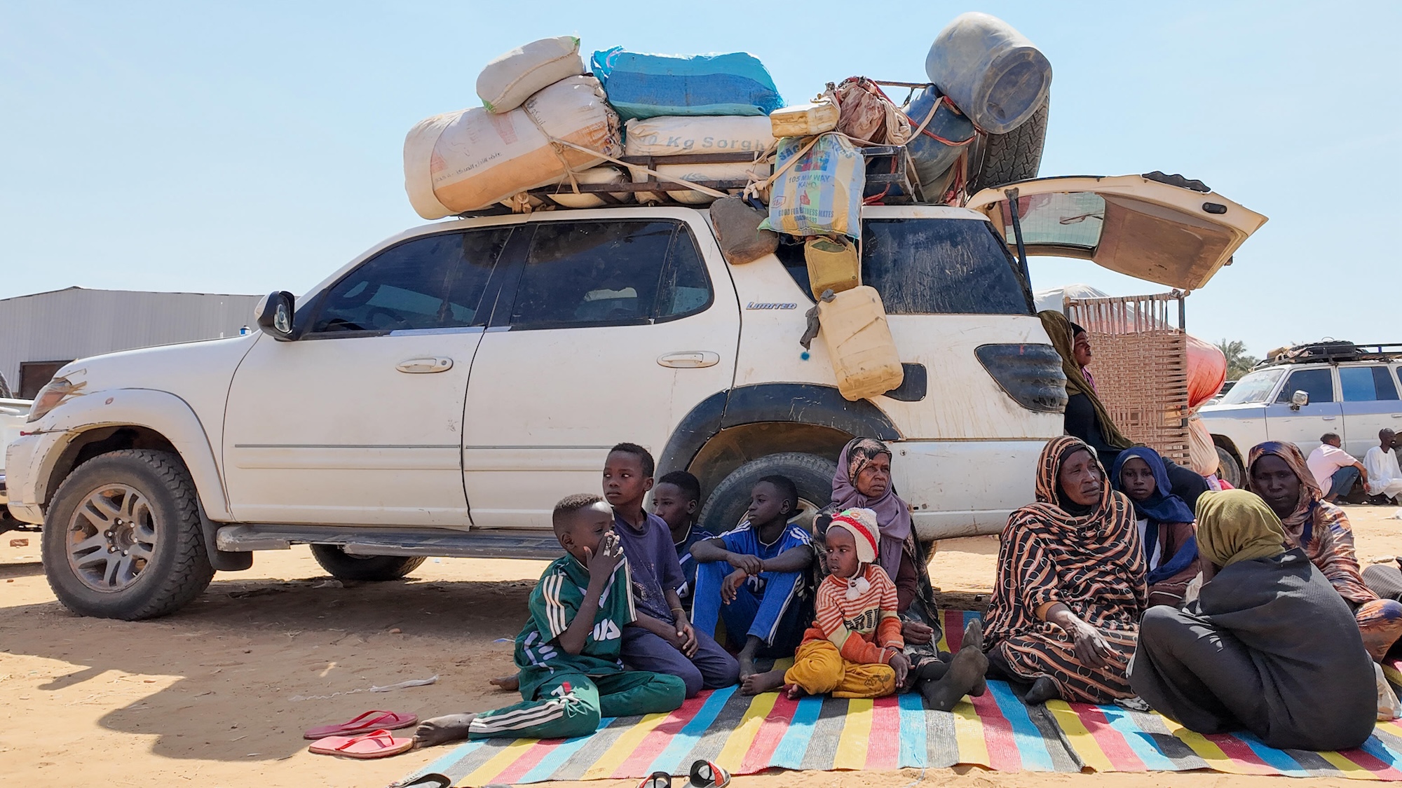 A family sit on a blanket on the ground in front of a truck loaded with bags and suitcases.