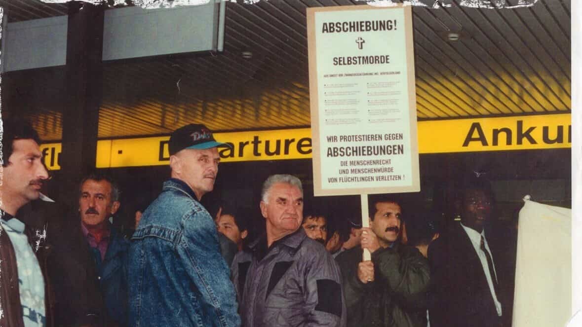 Männer stehen in einer Flughafenhalle und halten ein Schild gegen Abschiebungen hoch.