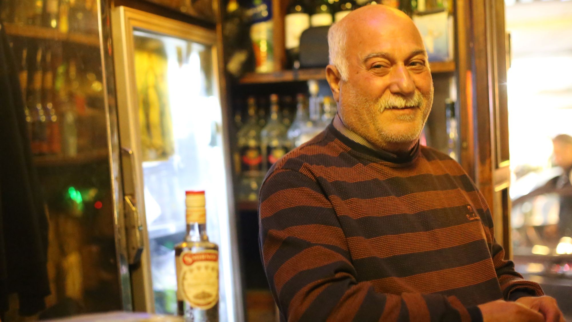 Ein Mann im gestreiften Pullover, mit weißem Schnauzbart und Haar, steht in einer Bar.