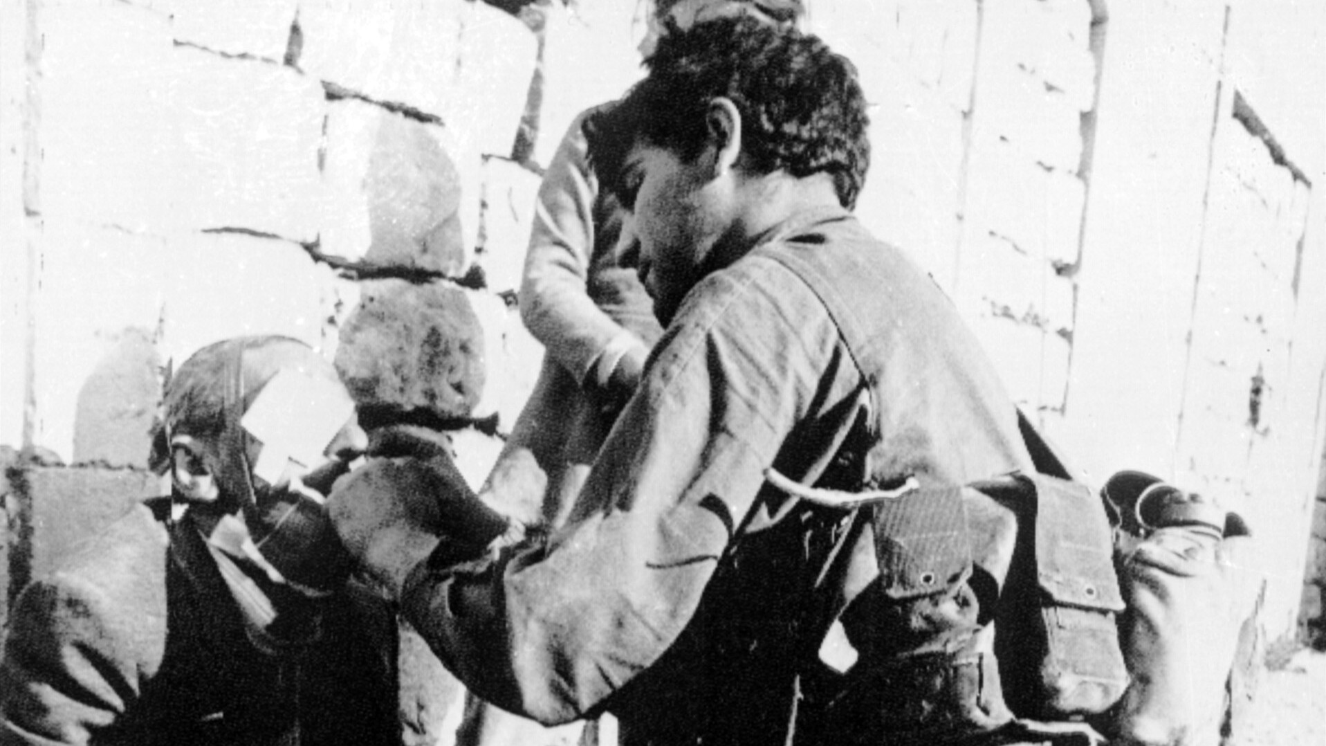 Schwarz-weiß Foto: Ein Soldat behandelt einen verletzten alten Mann, der an eine Mauer gelehnt sitzt.