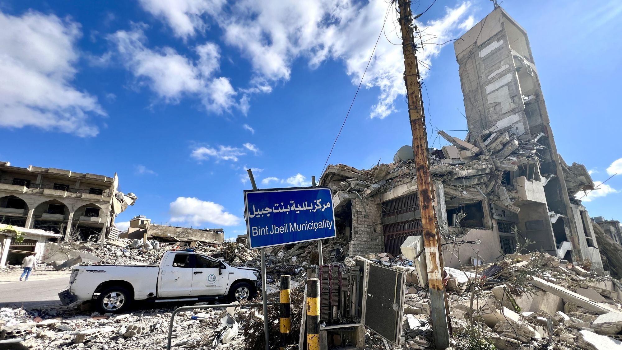 Im Vordergrund ein Schild mit der Aufschrift "Bint Jebeil Muicipality", dahinter Trümmer und ein Pick-Up Truck