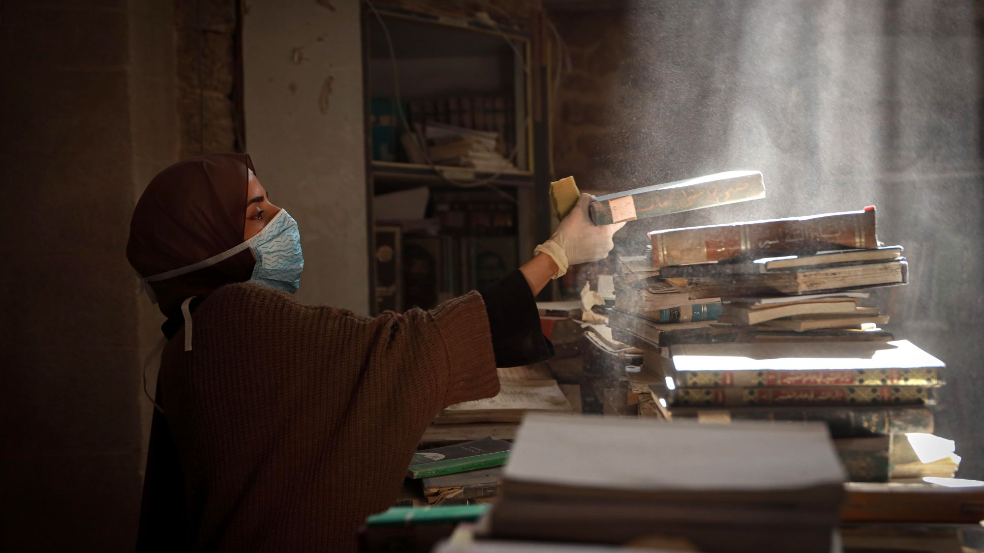 A woman wearing a hijab and a face mask dusts books in a destroyed library.