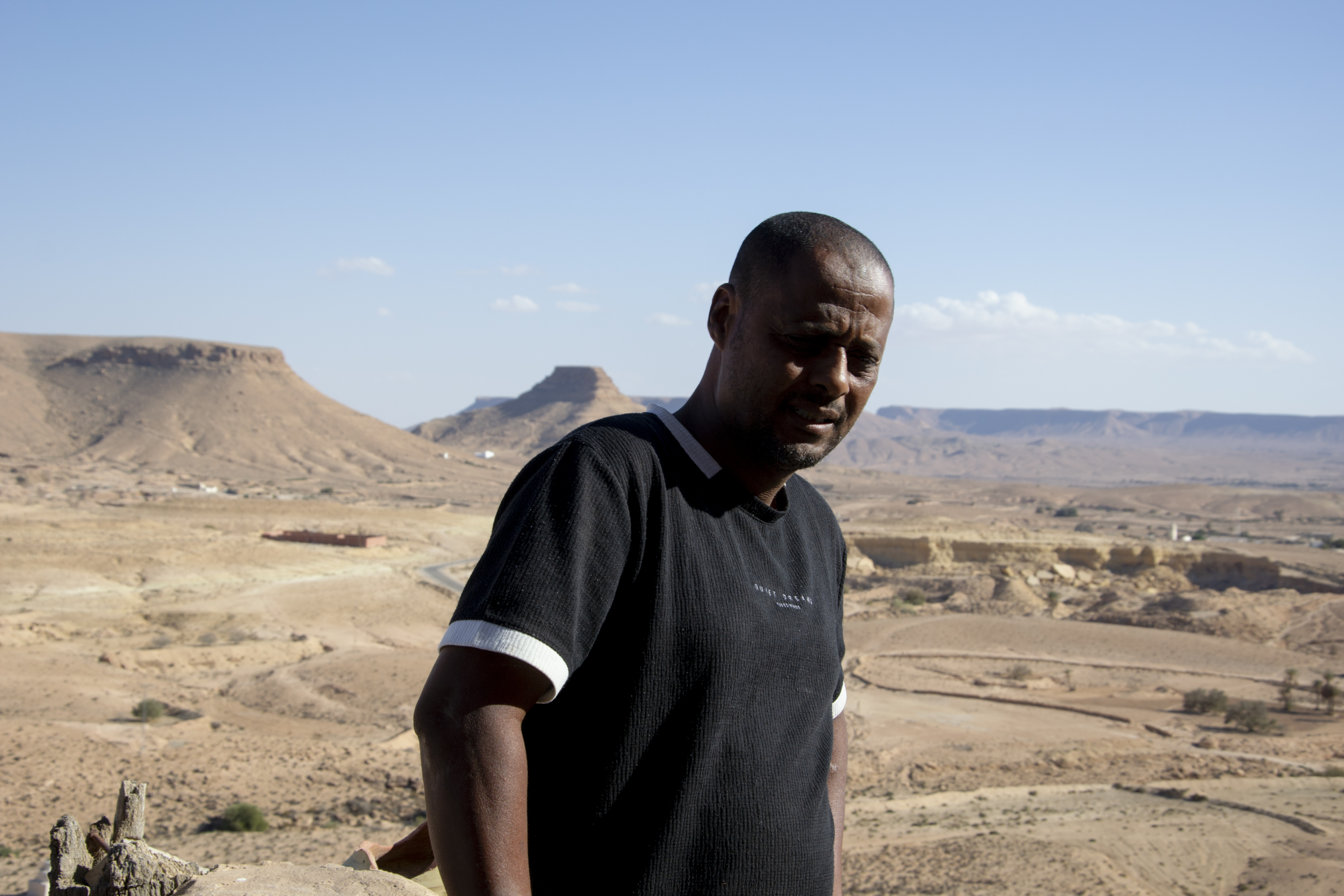 A man stands in front of a vast desert landscape. Mountains visible in the background.