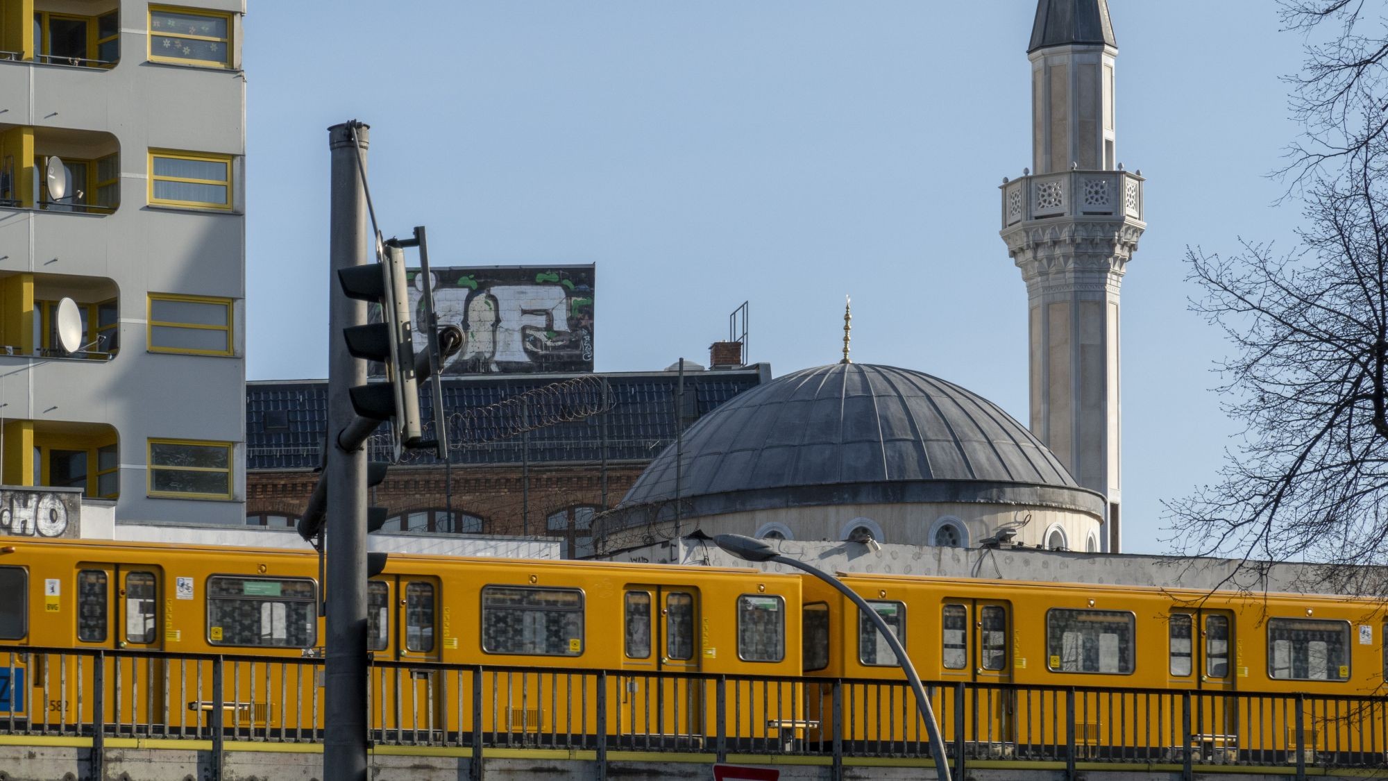 Stadtbild, im Vordergrund fährt eine gelbe Bahn, im Hintergrund ein Wohngebäude und eine Moschee.
