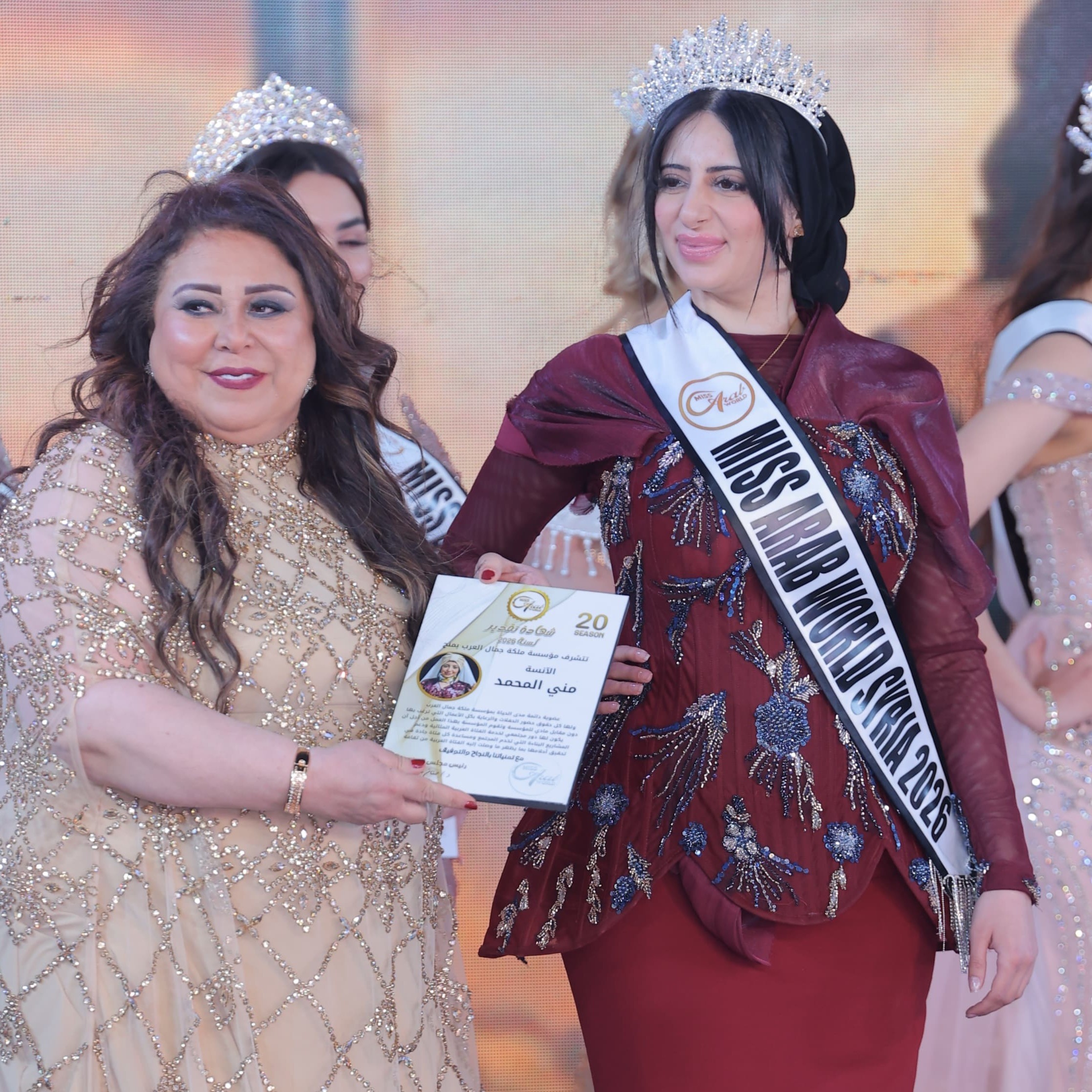 zwei Frauen in festlicher Kleidung, die ältere hält eine Urkunde in der Hand. Die jüngere hat eine Krone auf dem Kopf und trägt eine Schärpe mit der Aufschrift "Miss Arab World Syria 2026"