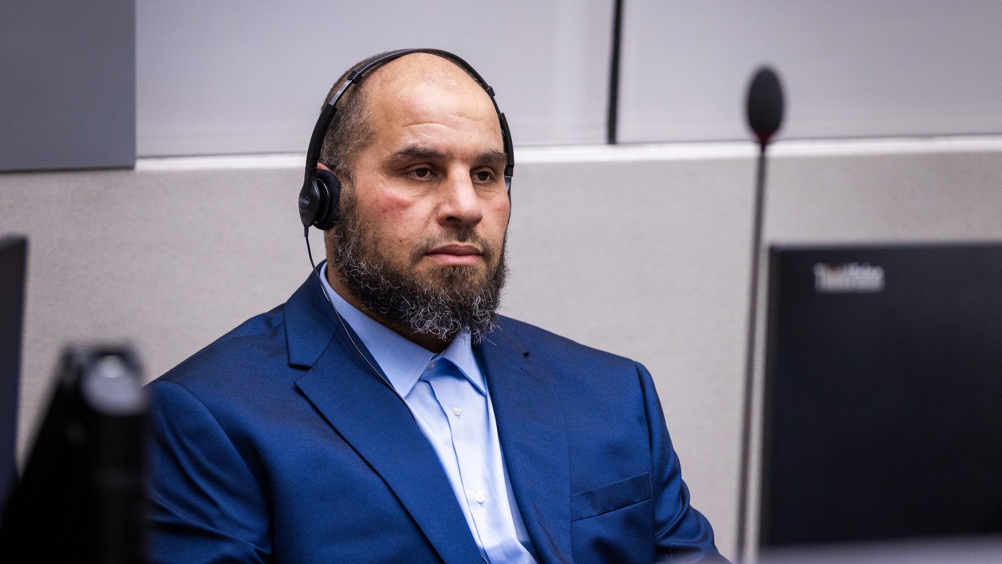 A man with a beard and headphones, wearing a suit, is sitting in a courtroom.