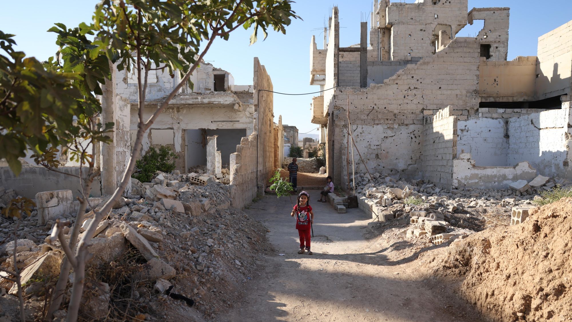 A girl is standing on a path between dilapidated buildings and rubble. In the background are another girl and an old man.
