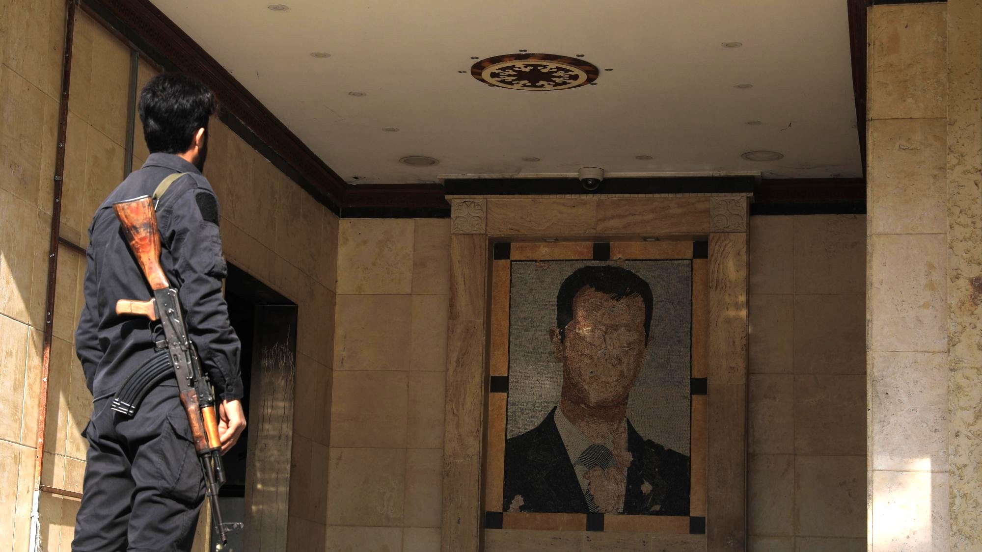 A man in uniform, holding a rifle, seen from behind. He is looking at a damaged mural of Bashar al-Assad.