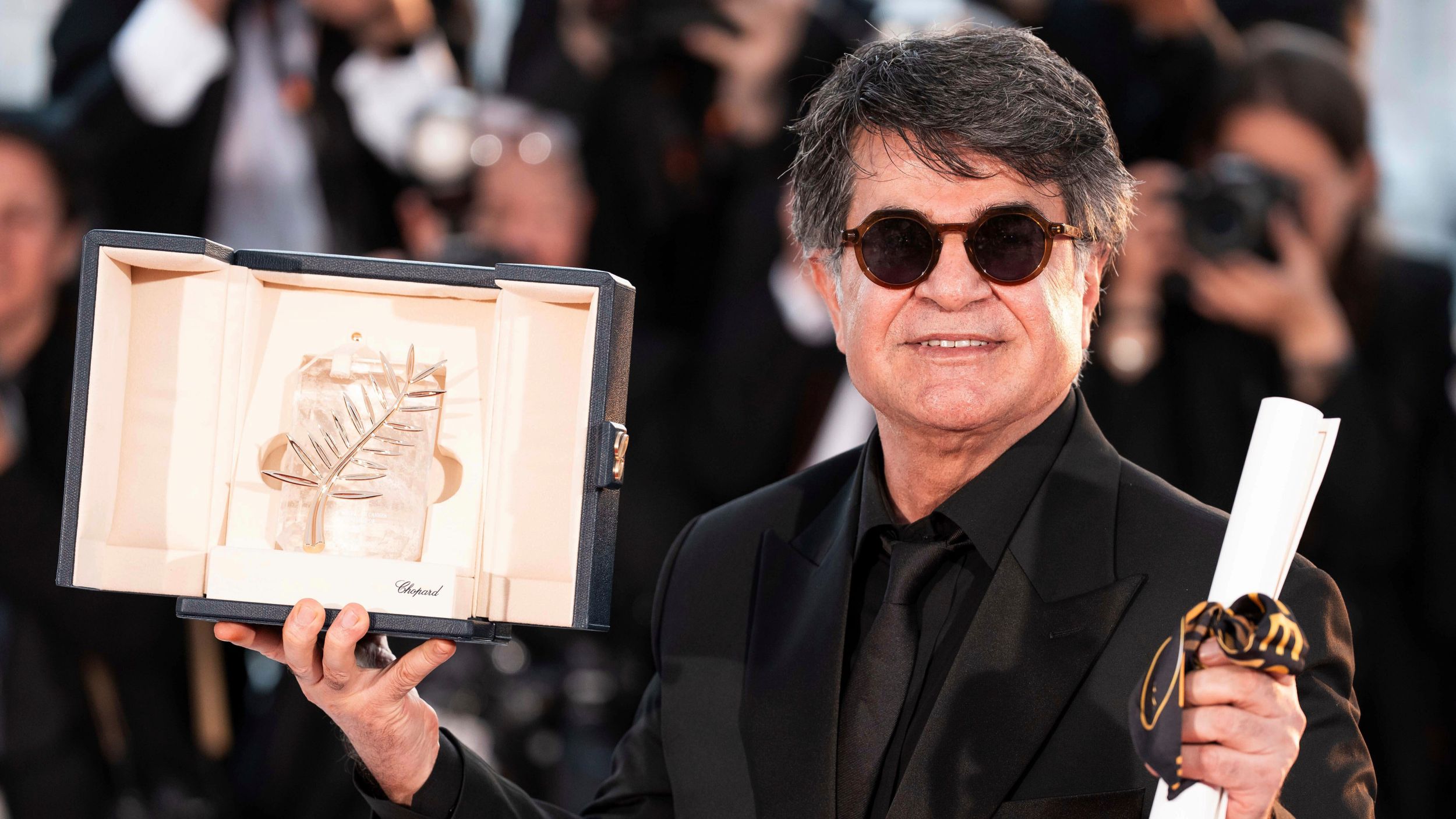A man wearing a black suit and sunglasses holds an award and a scroll.