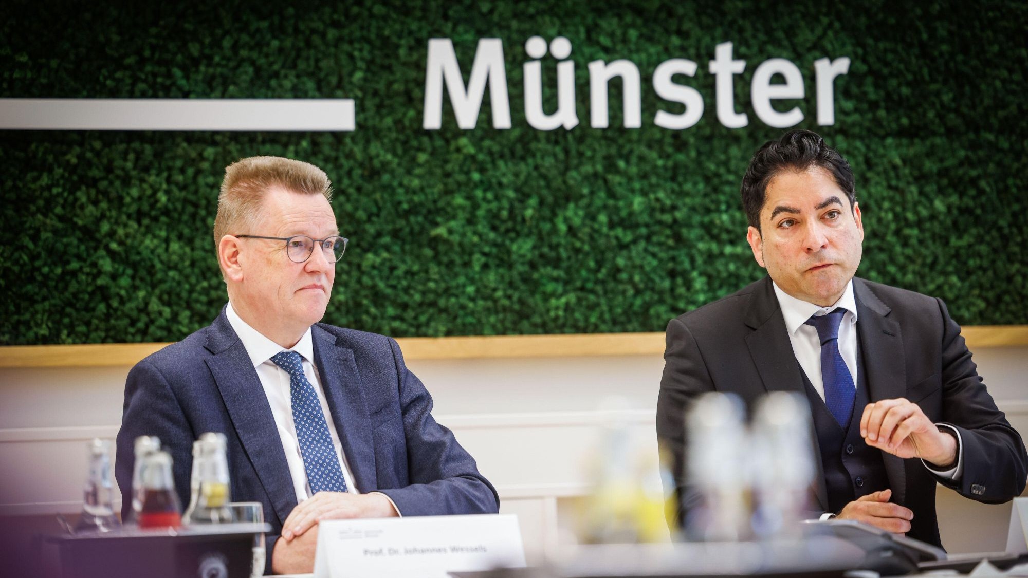 Zwei Männer in Anzug sitzen an einem Konferenztisch, im Hintergrund an der Wand der Schriftzug "Münster"