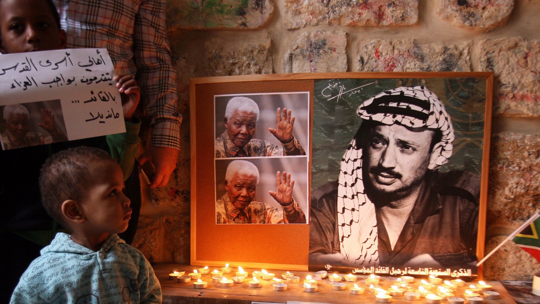 Two children stand beside a board holding portraits of Nelson Mandela and Yasser Arafat. Below the portraits are candles.