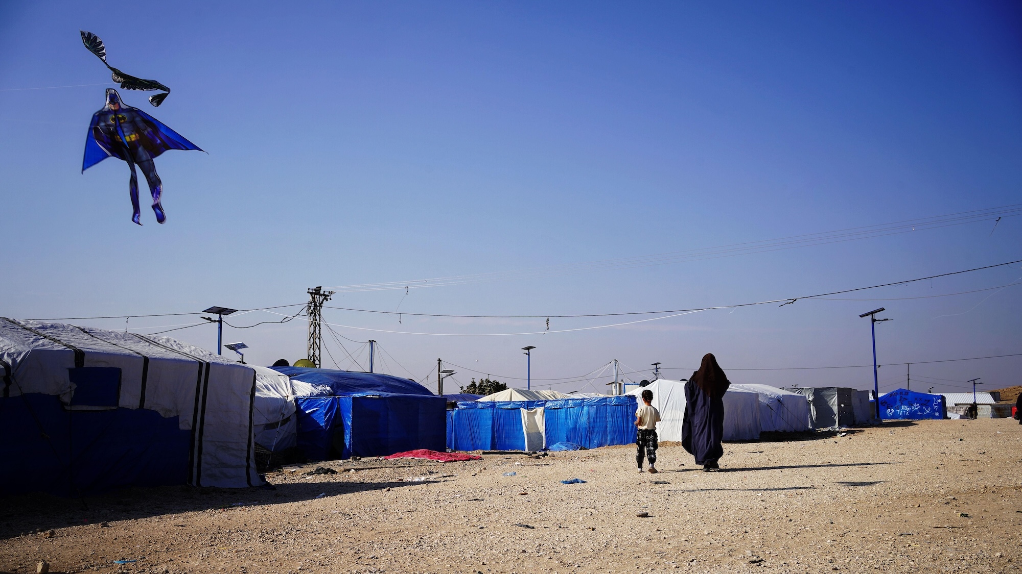 A woman and a child walk towards a cluster of tents. A Batman shaped kite is visible in the sky.