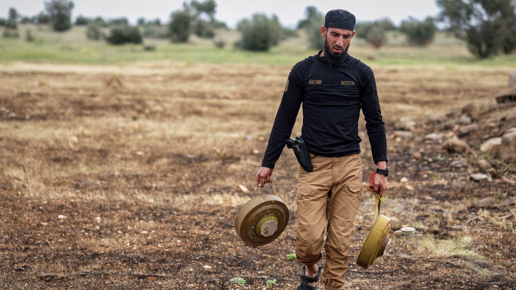 A man walks through a field carrying two large circular mines.