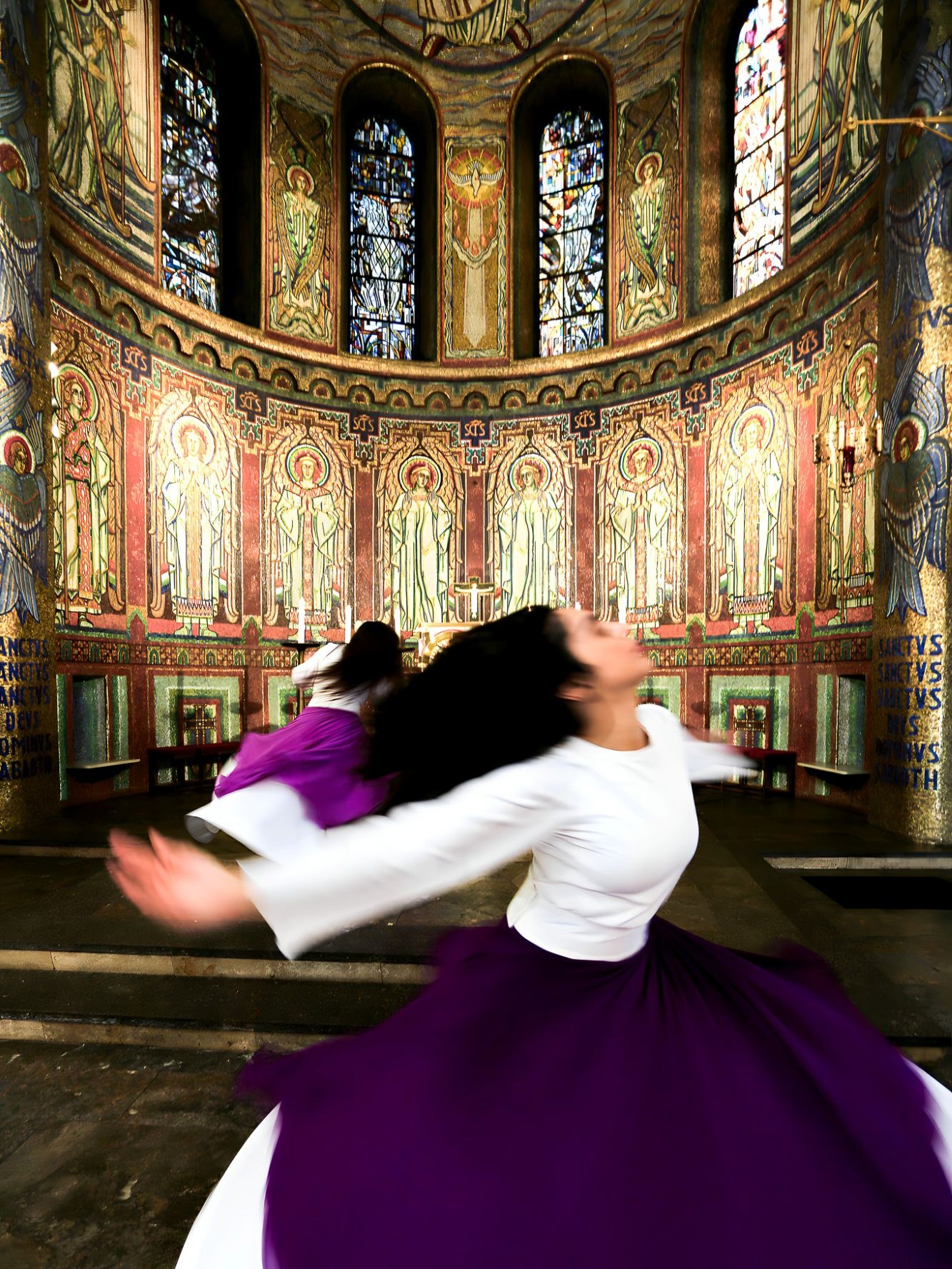 Two women in white robes and purple aprons are dancing in a festively lit church.